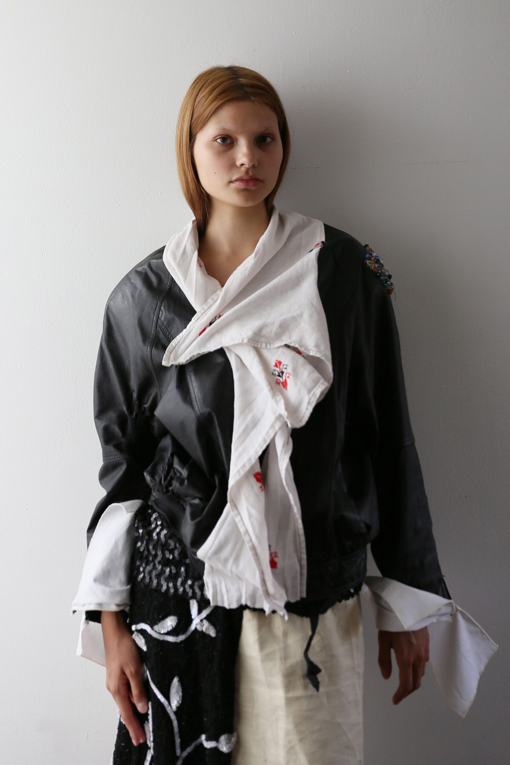 Black leather jacket cinched at the waist, one shoulder crusted with beads. White cotton shirt twisted into a draped scarf front, cuffs hanging long. Patchwork skirt split between black sequins and raw ivory linen. After the opening, still wearing the tablecloth and the spill of confetti.