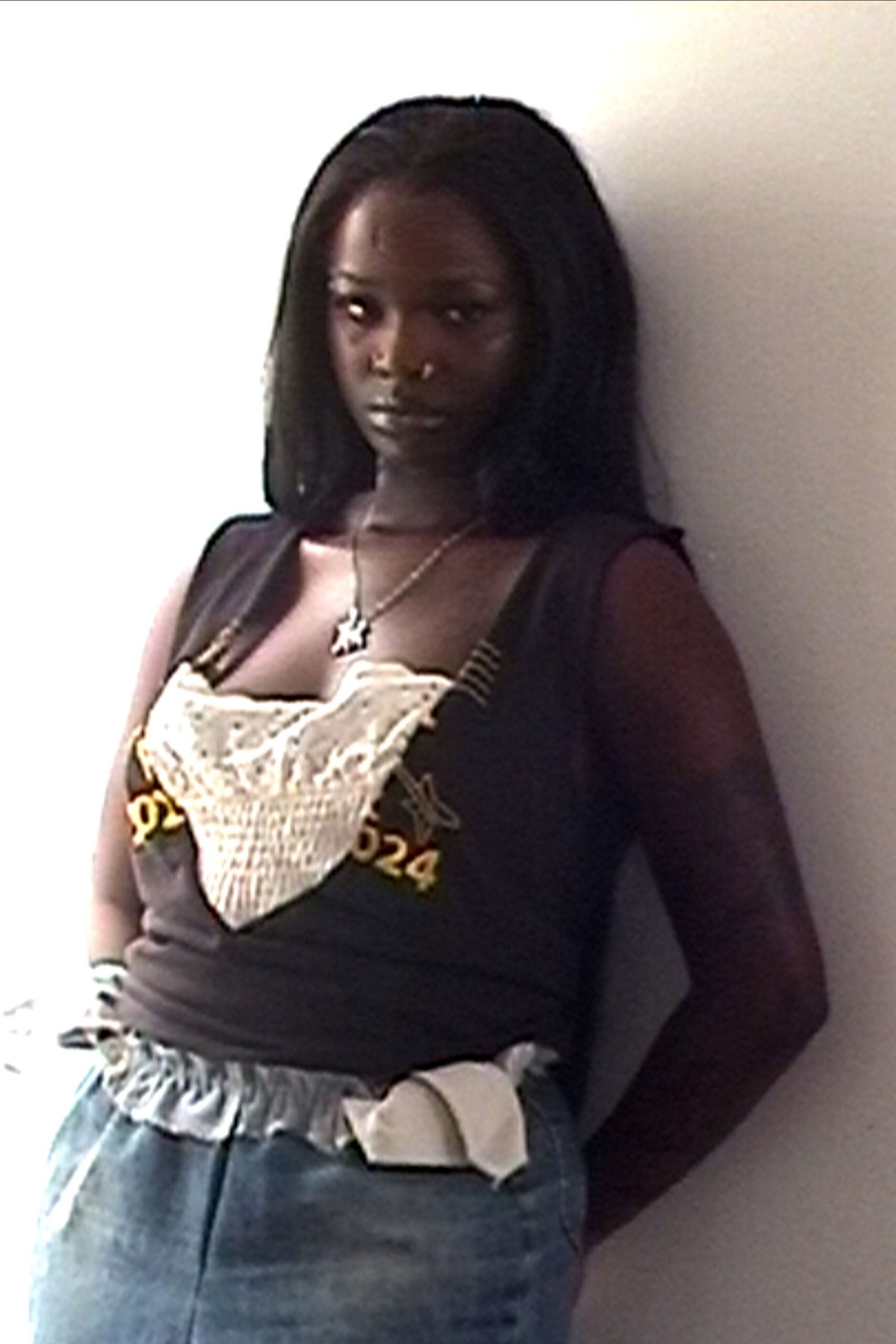 Silver pendant necklace hanging at the collarbone. Black fitted tank with yellow numerals and a cream crochet bib. Stacked striped bangles at one wrist. Loose blue denim with a gathered paperbag waist and white pocket lining. Leaning in a hot hallway after the house party emptied out.