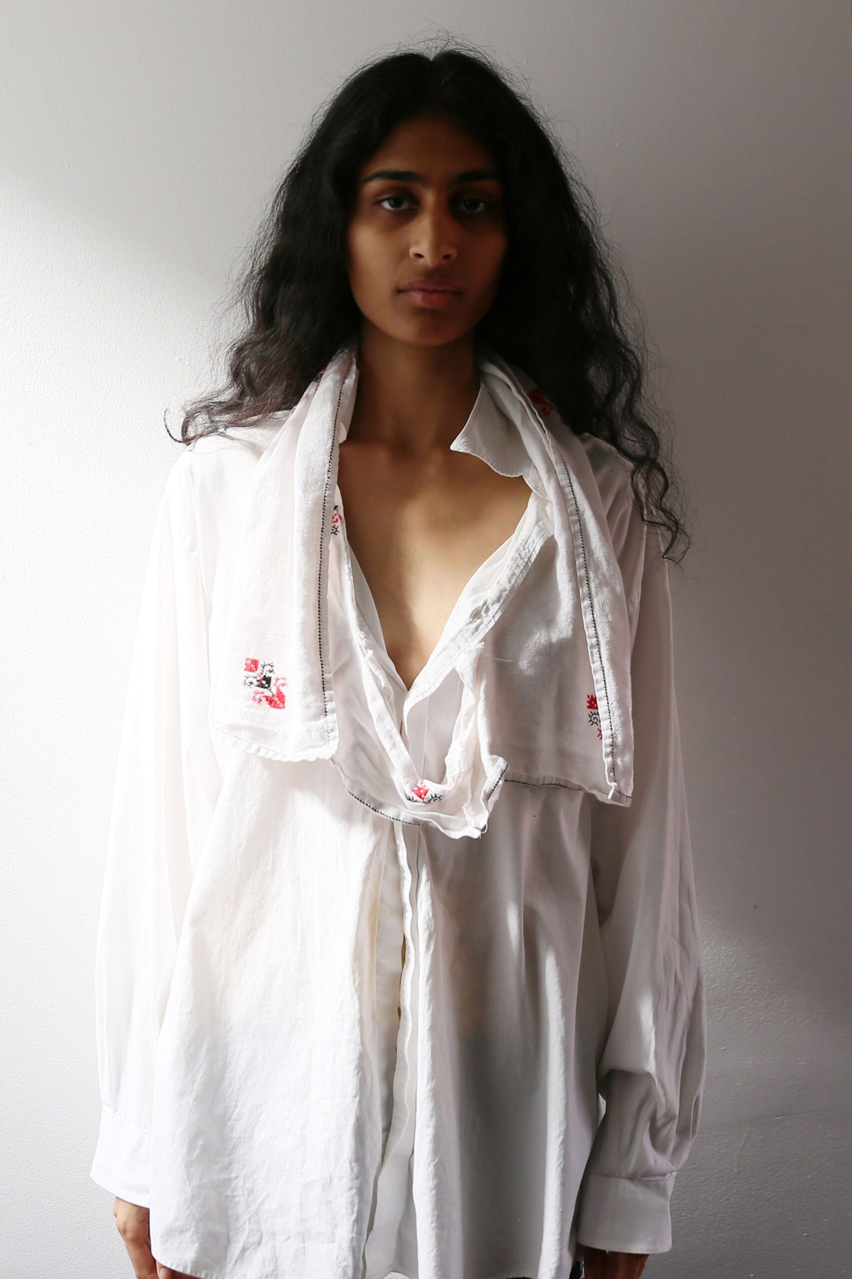 White cotton scarf with tiny red-and-black cross-stitch and dark whipstitched edges. Oversized white shirt in thin washed cotton, open deep at the chest. Morning light against apartment plaster, still wearing yesterday's embroidery.
