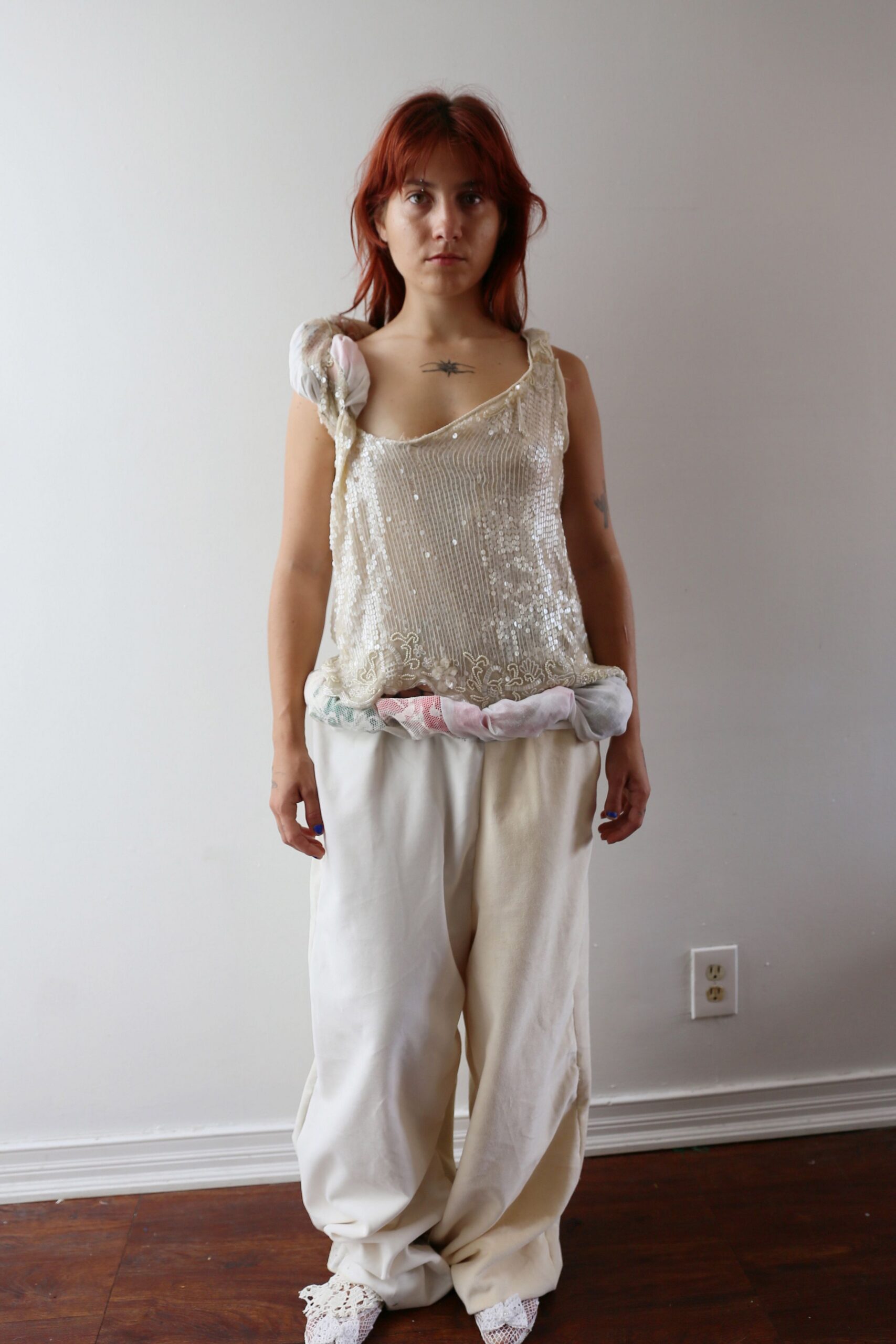 Ivory sequined tank, scalloped hem and beadwork slipping off one shoulder. Stuffed mesh tubes circling the hips in pale pink and mint. Cream wide-leg trousers, pooling into soft folds at the ankles. White lace-covered pointed boots. Morning after a wedding, standing barefoot in someone else's dining room.