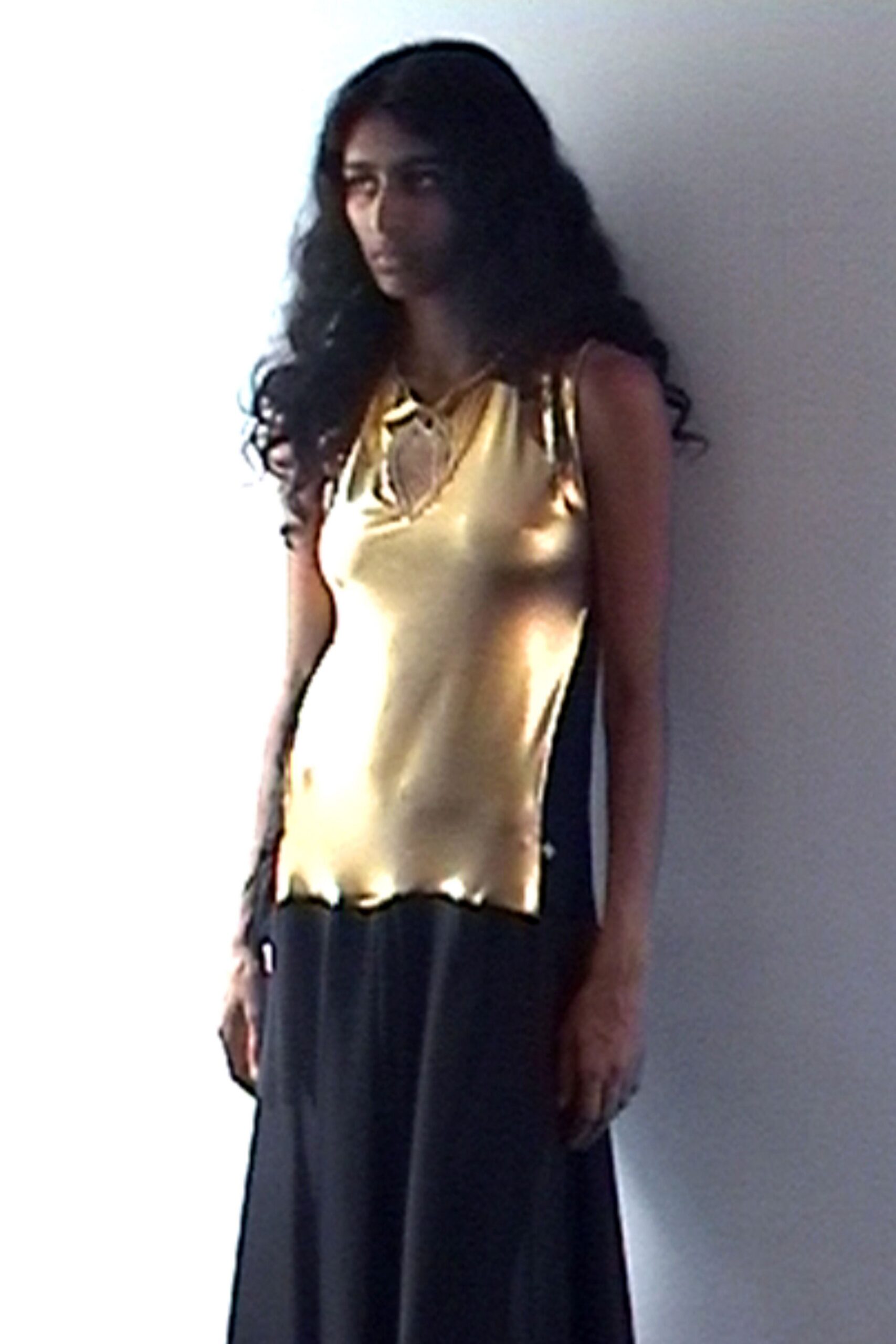 Liquid gold satin tank, teardrop keyhole tied with a narrow cord. Black floor-length skirt hanging straight from a dropped seam. Slim black shoulder bag tucked behind the arm. Afternoon light in a half-empty apartment before the mirror is hung.