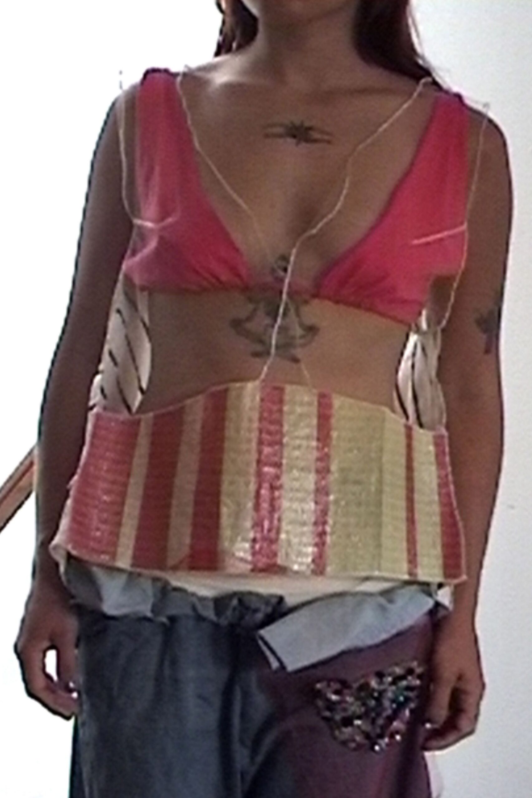 Hot-pink satin bralette, gathered cups tied together at the sternum. Cream and rose striped woven panel top, raw cords tracing the bare torso. Slouchy two-tone trousers with a pale chambray sash knotted low. Beaded butterfly patch glittering on the upper thigh. Heat-hazed boardwalk afternoon, salt drying white on your shoulders.