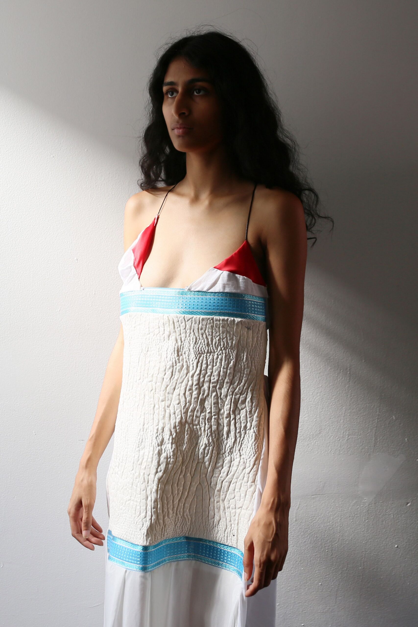White slip dress with red satin cups, black spaghetti straps, and woven aqua bands. Quilted ivory panel hangs straight from the bust, puckered like crushed bark. Sunlit hallway after swimming, towel marks still pressed into the skin.