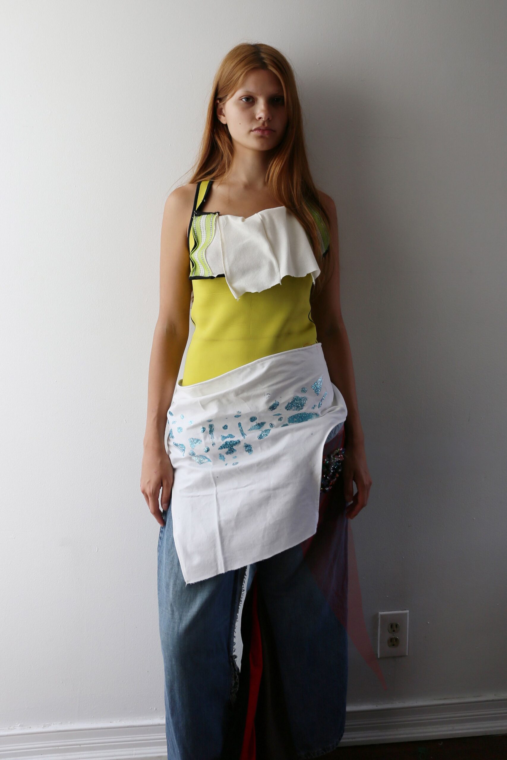 Acid-lime fitted top with black binding and a drooping white bib panel. White cotton apron wrap slung low, scattered with turquoise glitter marks. Wide blue denim legs split by a raw inner seam and red inset. Waiting by the studio wall after somebody spilled craft glitter on lunch.
