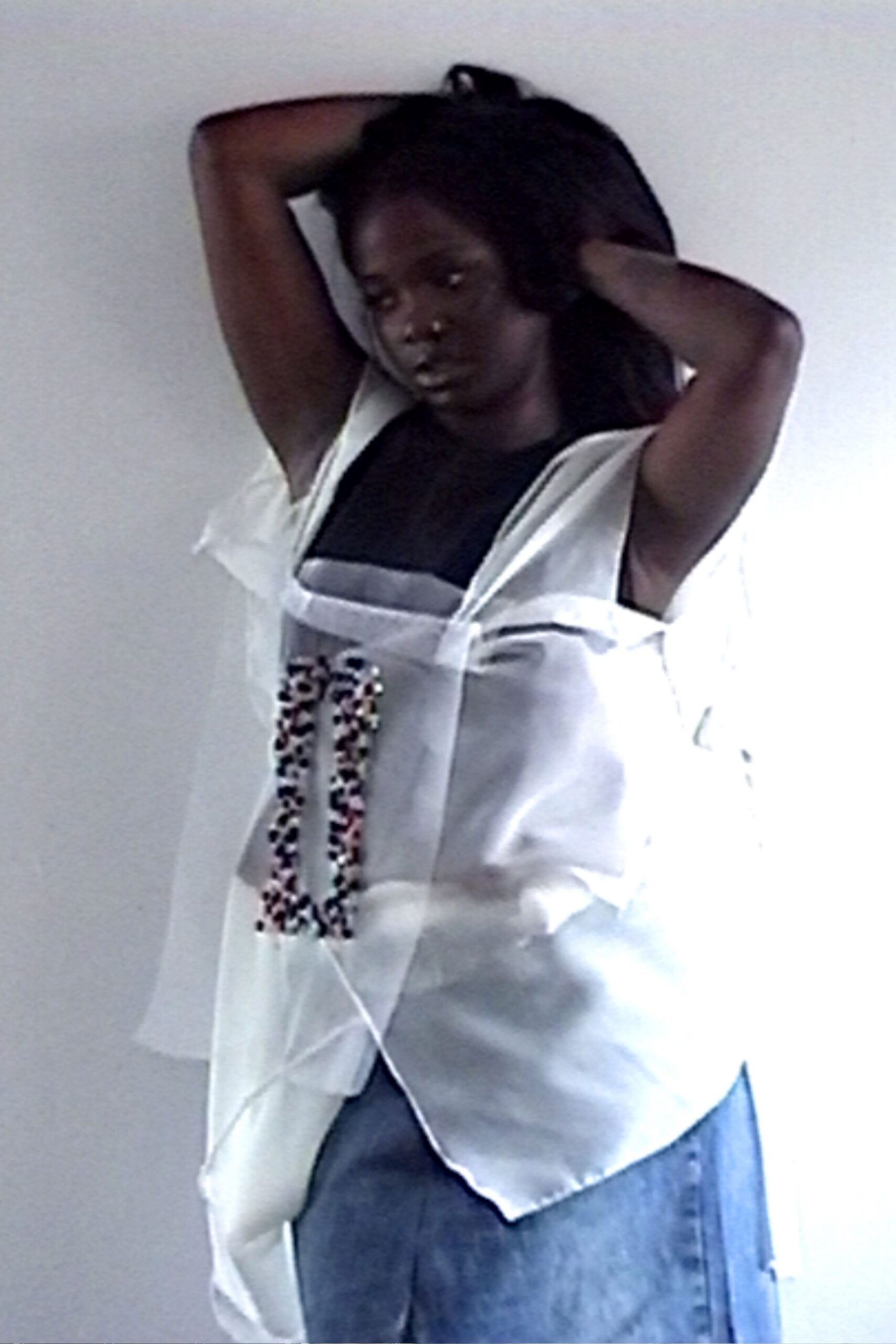 White satin apron top with loose side panels and a beaded U. Black jersey tank filling the square neckline. Washed denim hanging straight beneath the uneven satin hem. Afternoon in a borrowed studio, one window blowing everything sideways.