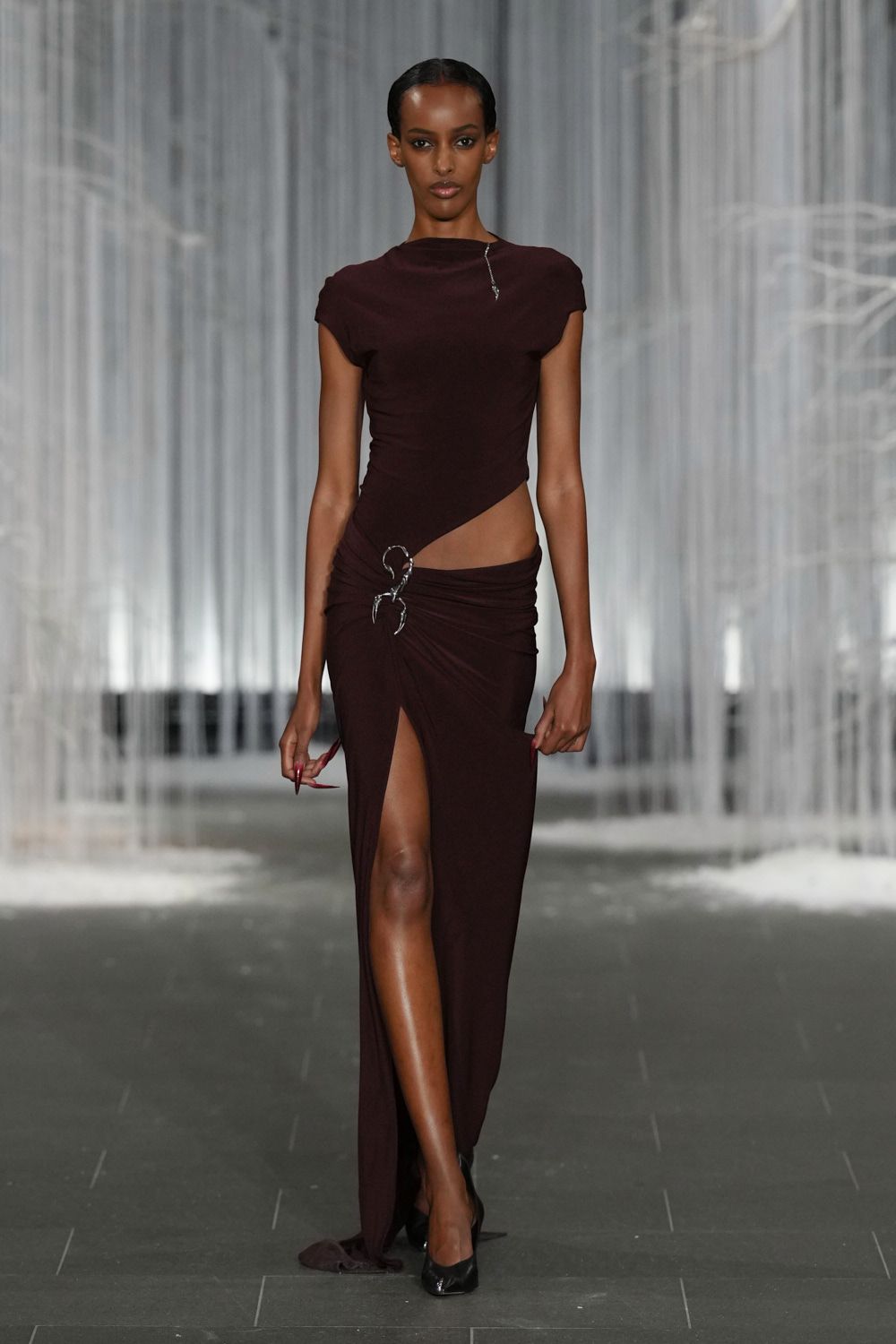 Dark aubergine jersey top with cap sleeves and a skewed cutout waist. Matching draped jersey skirt, thigh slit pinned by a silver scorpion clasp. Black pointed pumps. Whatever you wear to cross a hotel lobby after midnight in winter.