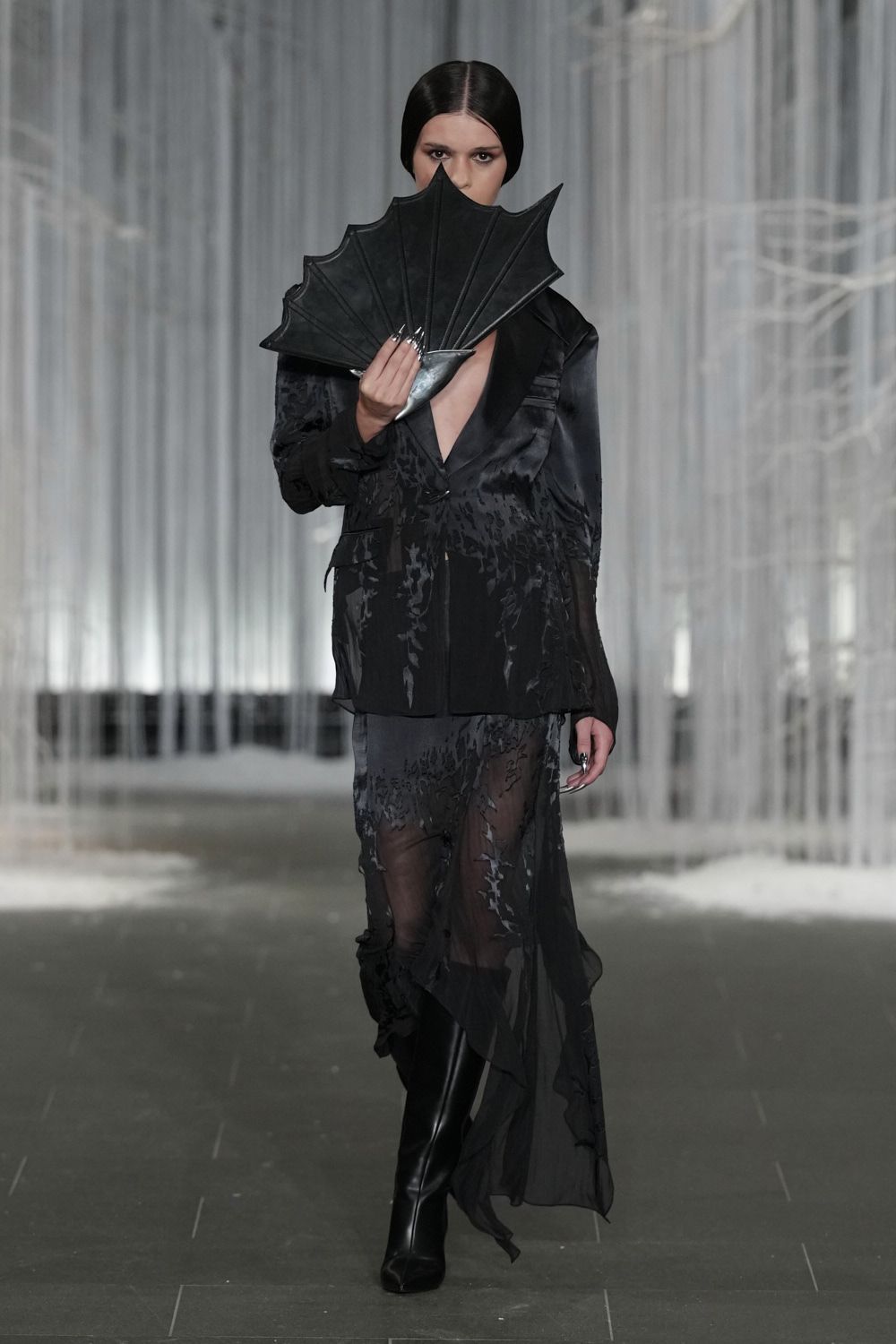 Black leather folding fan with batwing ribs held over the mouth. Inky devore blazer dress, satin shawl lapels plunging to the sternum. Sheer uneven hem drifting from the blazer in scorched floral velvet. Black knee boots with a hard square toe. Crossing a hotel atrium after midnight while the fountain is shut off.