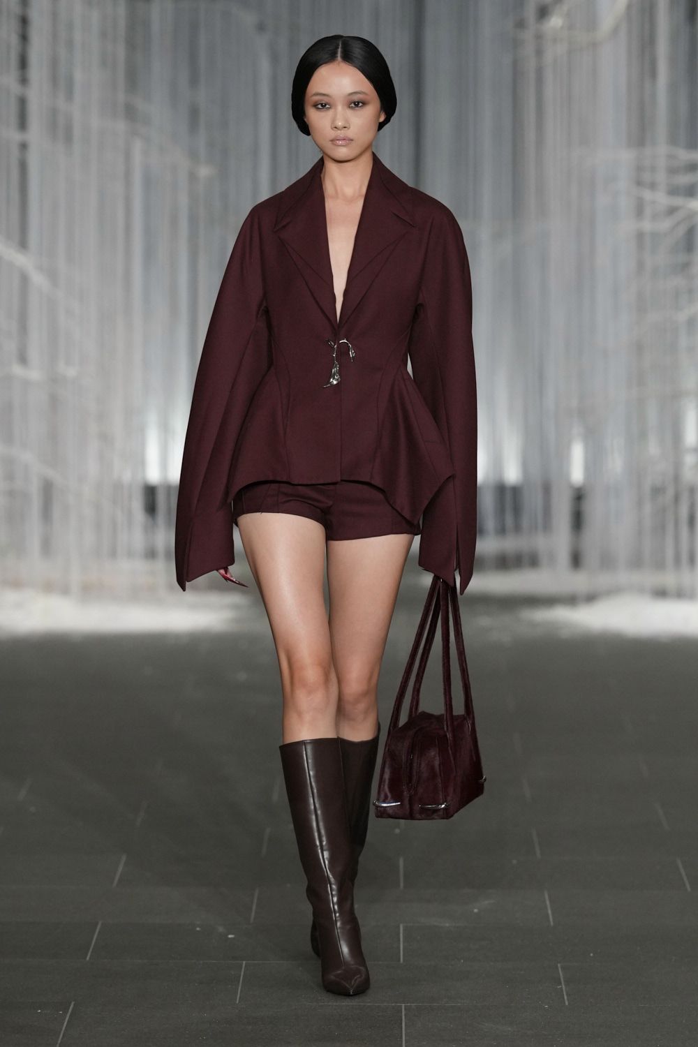 Oxblood blazer with sculpted hips and a tiny silver clasp. Matching tailored shorts cut high on the thigh. Plum suede tote with long straps and metal-capped corners. Dark brown leather knee boots with squared, inward-turning toes. Crossing an empty hotel atrium before anyone else is downstairs.