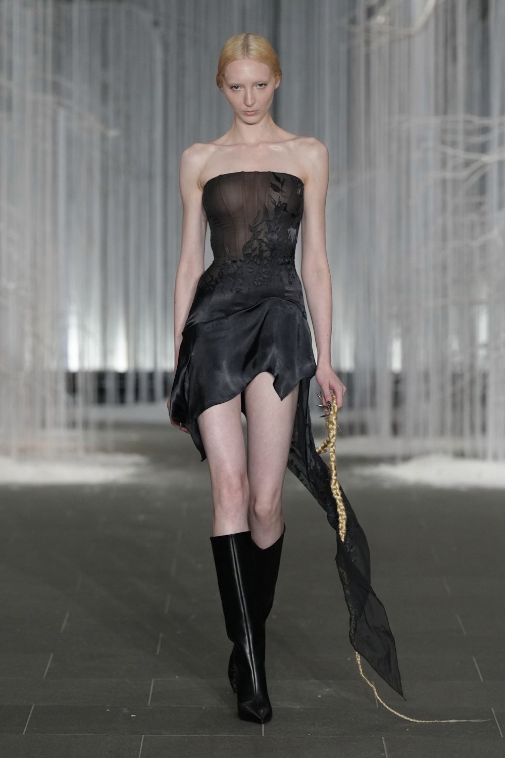 Black strapless dress with a sheer corseted bodice and scorched floral appliqué. Asymmetric satin skirt melting into a long gauze panel. Knife-point black leather boots to the knee. Thick blond braid looped from the fingers, trailing a gold cord end. Crossing a museum atrium after midnight with something strange still in hand.