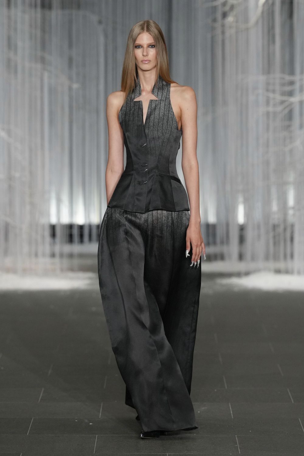 Charcoal halter top with pinstriped bib and a sharp notch at center chest. Matching satin wide-legs, cut long enough to drag over the shoe. Black pointed toes barely surfacing under the hem. Hotel ballroom after midnight, stacking banquet chairs while the band packs up.