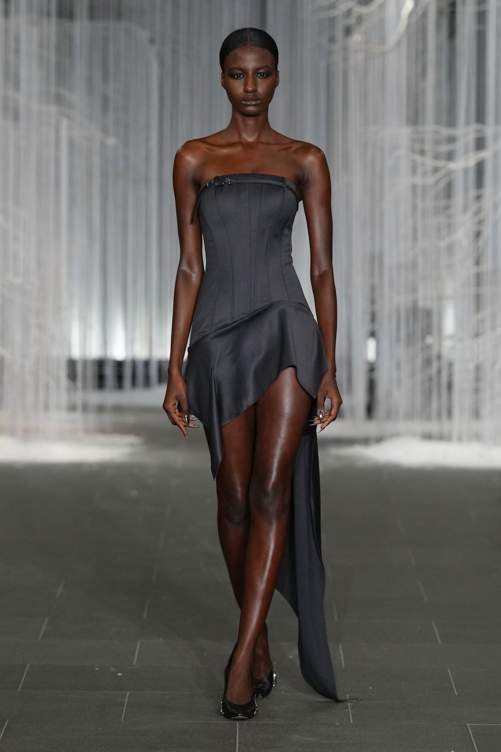 Charcoal strapless corset dress with channel seams and a tiny buckle tab. Black satin drape cut high on one thigh, falling to the floor. Black pumps with a silver ring glinting at the heel. Whatever you wear to cross a hotel lobby after midnight without breaking stride.