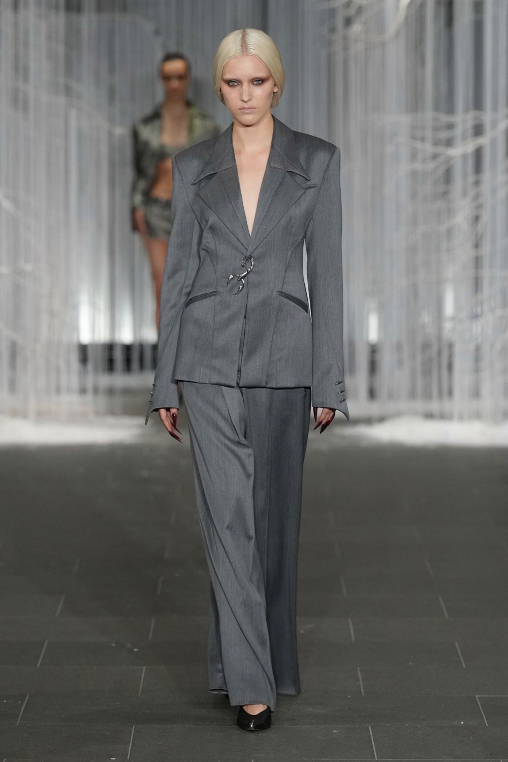 Grey worsted blazer with blade shoulders and a silver scorpion clasp. Matching wide-leg trousers breaking softly over pointed black pumps. Patent black pumps, squared off at the vamp. For crossing a hotel lobby at dawn before anyone from the conference is up.