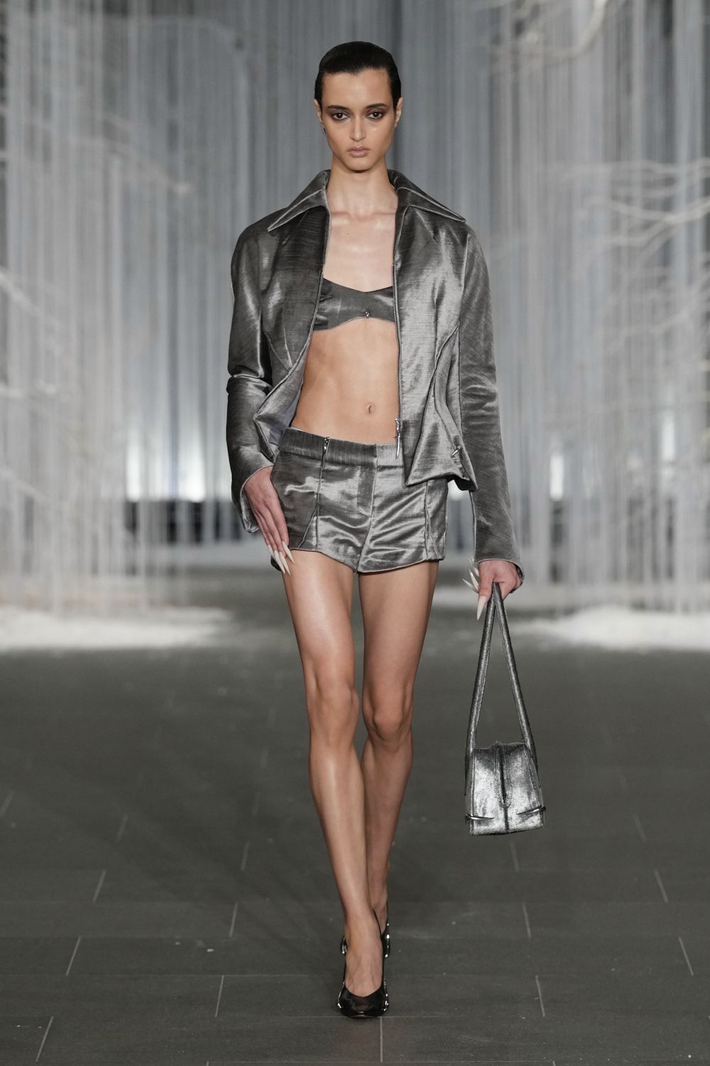 Gunmetal lame jacket with a wide collar and low zip pull. Matching bandeau bralette, tiny clasp glinting at the center. Low-rise metallic shorts with pressed seams and curved split hems. Slouchy silver tote with long narrow straps. Black patent pumps. Crossing a skybridge after midnight with the office lights still on.