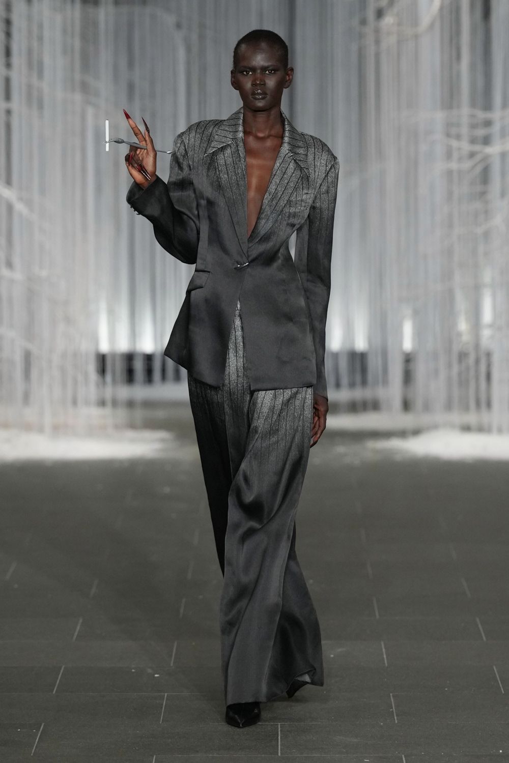Charcoal pinstriped blazer cut with peaked lapels and one low hook closure. Matching fluid pinstripe trousers, pooling lightly over the pointed shoe. Black pointed pumps almost hidden under the hem. Waiting for the tribunal doors to open after rain.
