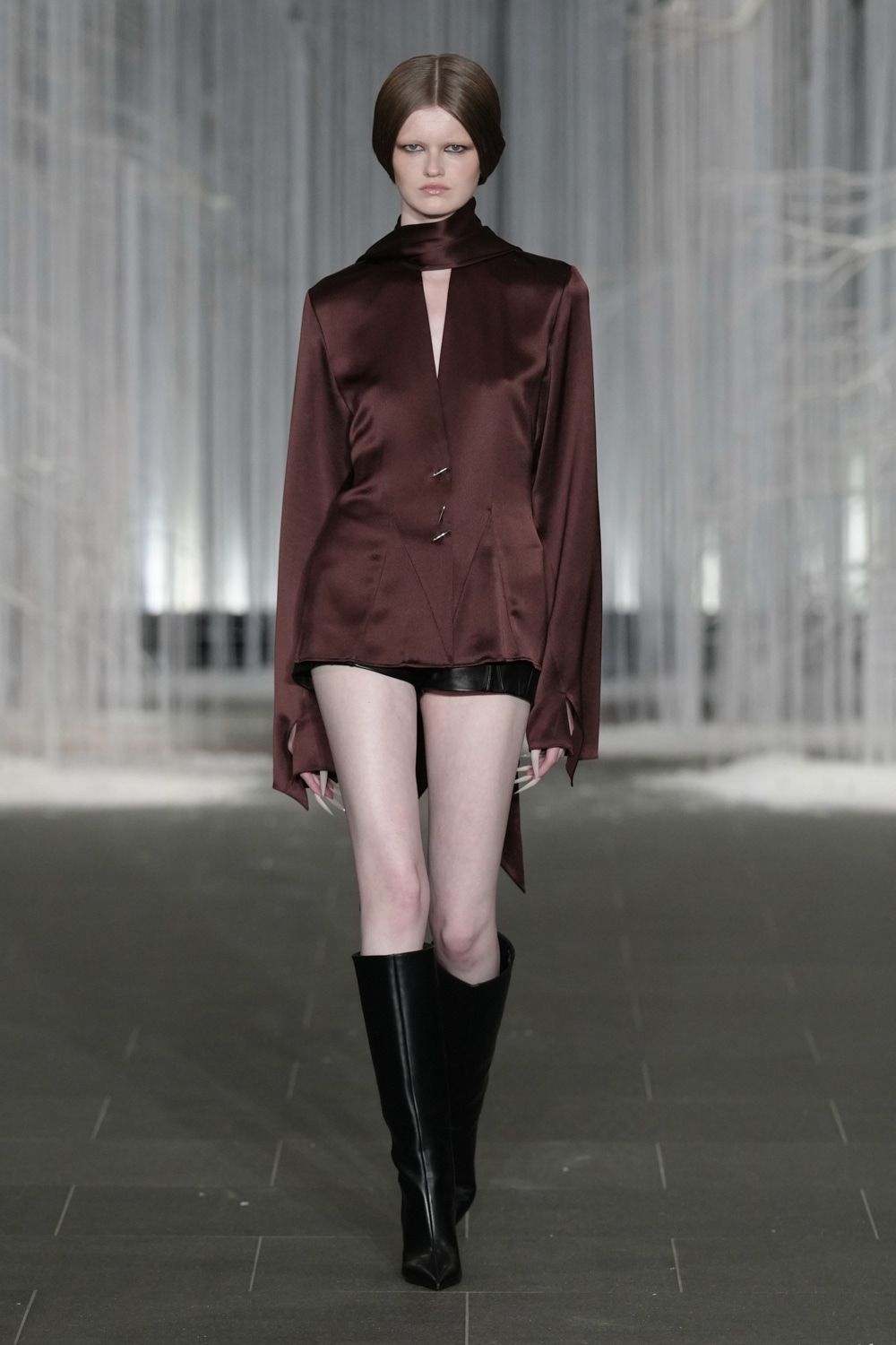 Oxblood satin blouse with a built-in scarf collar and dagger cuffs. Black leather hot pants, just a narrow strip under the hem. Black leather knee boots with hard squared toes. For crossing a hotel atrium after midnight with your room key missing.