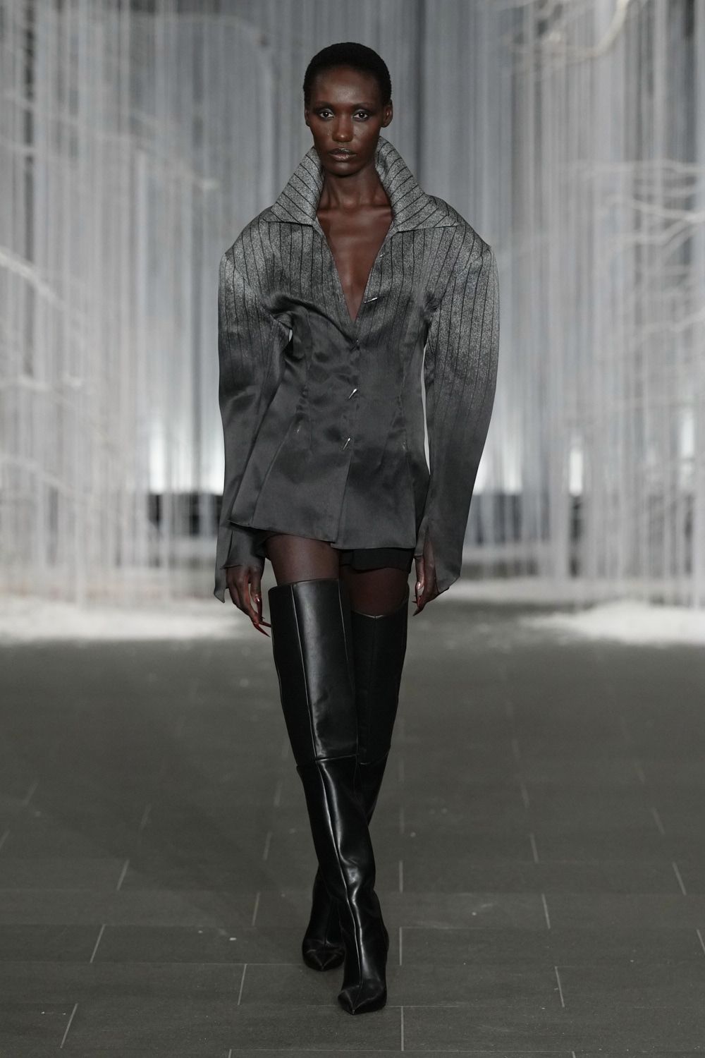 Smoke-grey satin blouse, pinstriped yoke rising into a wide funnel collar. Black shorts barely skimming below the blouse hem. Black leather thigh-highs with squared shafts and pinched pointed toes. Crossing a hotel atrium before dawn, heels ticking louder than the sprinklers.