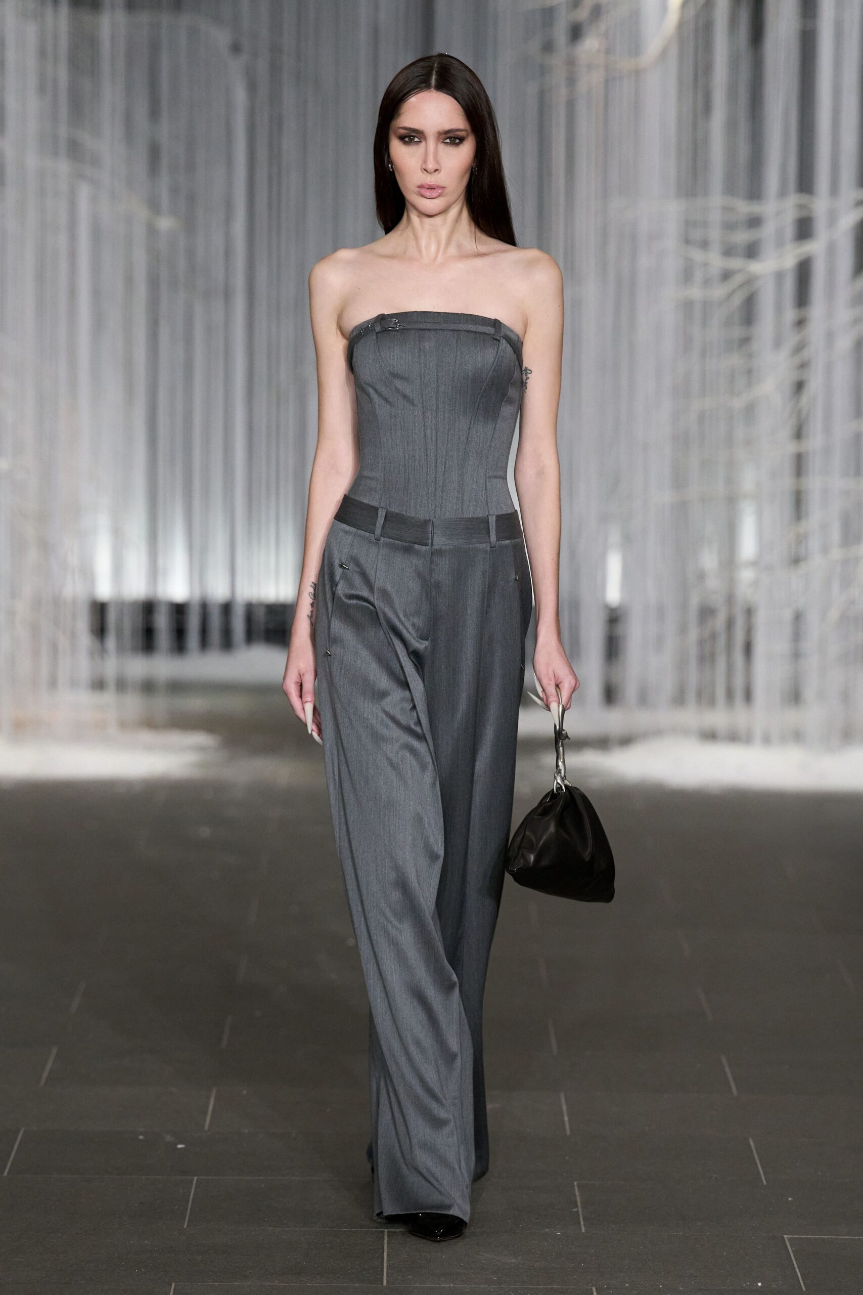 Strapless charcoal suiting corset, pinched with a tiny buckle at the top edge. Low-slung grey trousers with a black waistband and loose pooled break. Soft black pouch bag on a twisted silver handle. Black pointed toes just breaking under the hem. For crossing a hotel lobby at dawn before anyone else is awake.