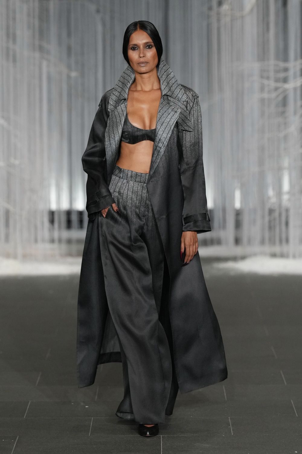 Floor-length charcoal coat with a giant pinstriped collar kicked upright. Black leather bralette, center seam and a tiny exposed zip. Wide black satin trousers with a pinstriped pleated waistband fold. Black pumps barely visible under the dragging hem. Whatever you wear to cross an atrium after midnight rain.