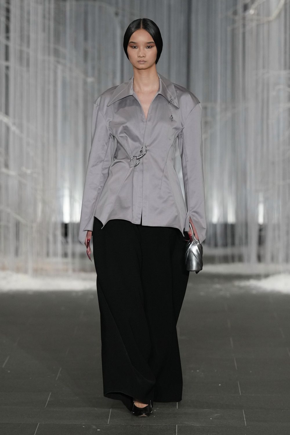 Pale grey fitted shirt in crisp satin, cinched with oversized silver safety pins. Black wide-legs falling in a dense, uninterrupted drape. Small silver pouch, gathered like a soft metal shell. Black patent square toes. Crossing a hotel atrium before anyone else comes down for breakfast.