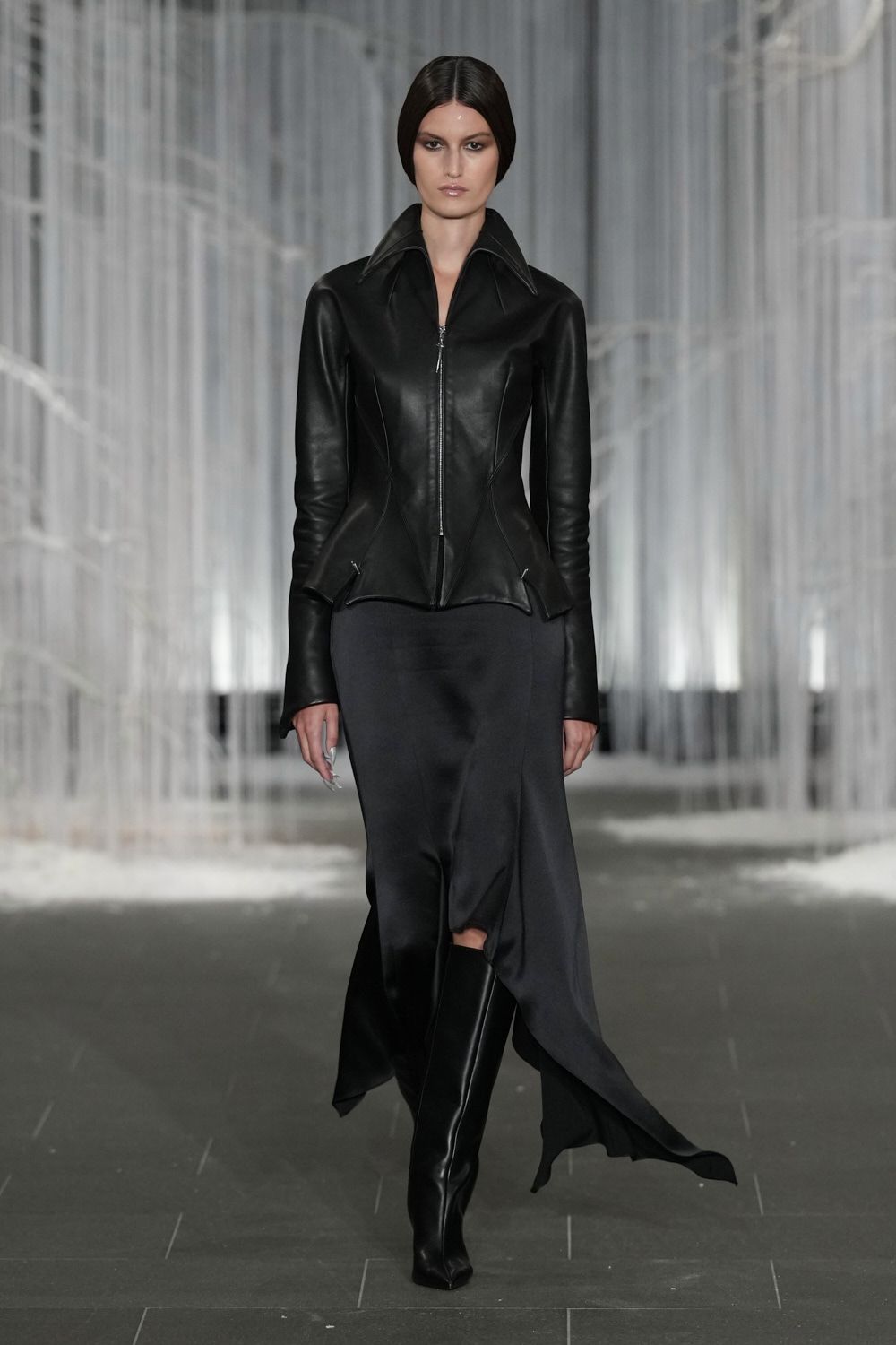 Black leather jacket, knife-sharp collar and seamed waist kicking out at the hips. Charcoal satin skirt cut on the bias, one hem tail dragging lower. Black leather boots with squared toes and a tight shaft. Crossing a near-empty hotel atrium before the conference breakfast starts.