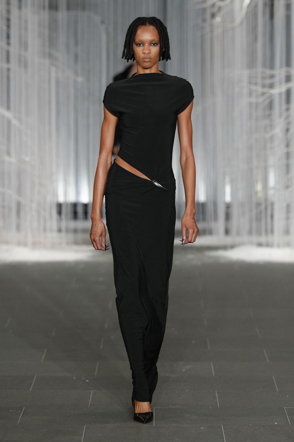 Black jersey top with a draped boatneck and cap-like sleeves. Matching column skirt cut with a slashed waist, pinned by an oval metal clasp. Black pointed pumps. Whatever you wear to meet someone under museum scaffolding after rain.