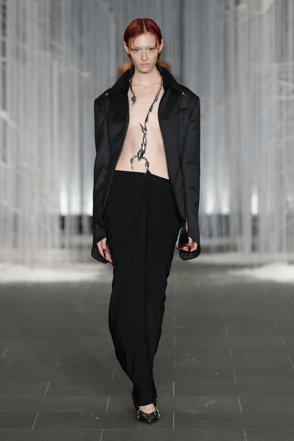 Black tailored jacket worn bare, collar spread wide and cuffs overlong. Chrome scorpion crawling from the neck to the waistband. Black jersey skirt knotted low, falling in heavy draped folds. Pointed black heels with a flash of silver at the toe. Crossing a marble hotel lobby after midnight with something venomous to say.