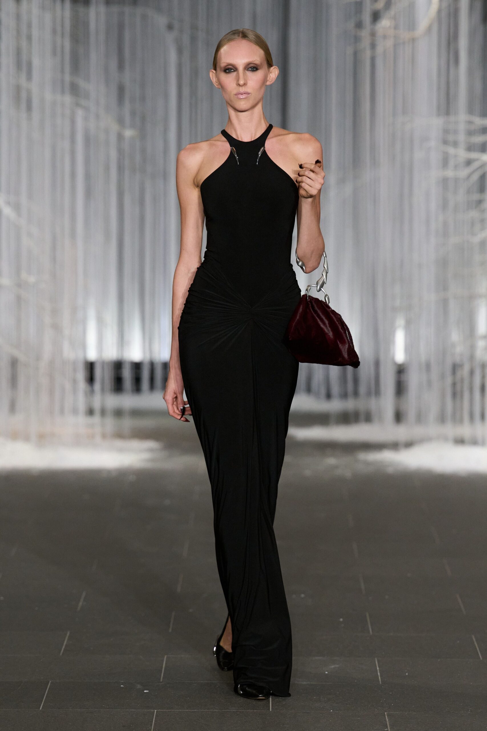 Black jersey halter gown, silver clips at the neckline, ruched into a low front twist. Burgundy satin pouch with a chunky linked silver handle. Black patent pumps peeking from the pooled hem. Crossing a hotel lobby after midnight with somebody else's room key.