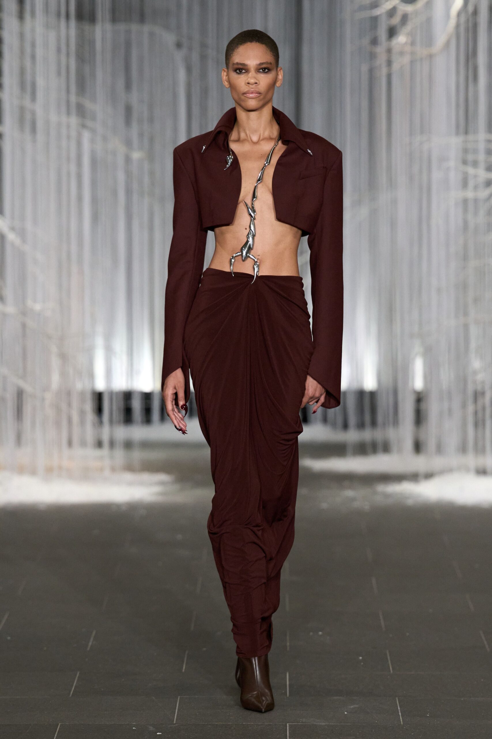 Oxblood cropped jacket with a wide collar and bare underbust cut. Twisted silver metal branch running from collarbone to low waist. Matching jersey skirt ruched into the harness at center front. Dark brown square-toe boots peeking from the pooled hem. Crossing a sculpture court just after rain, still cold off the stone.