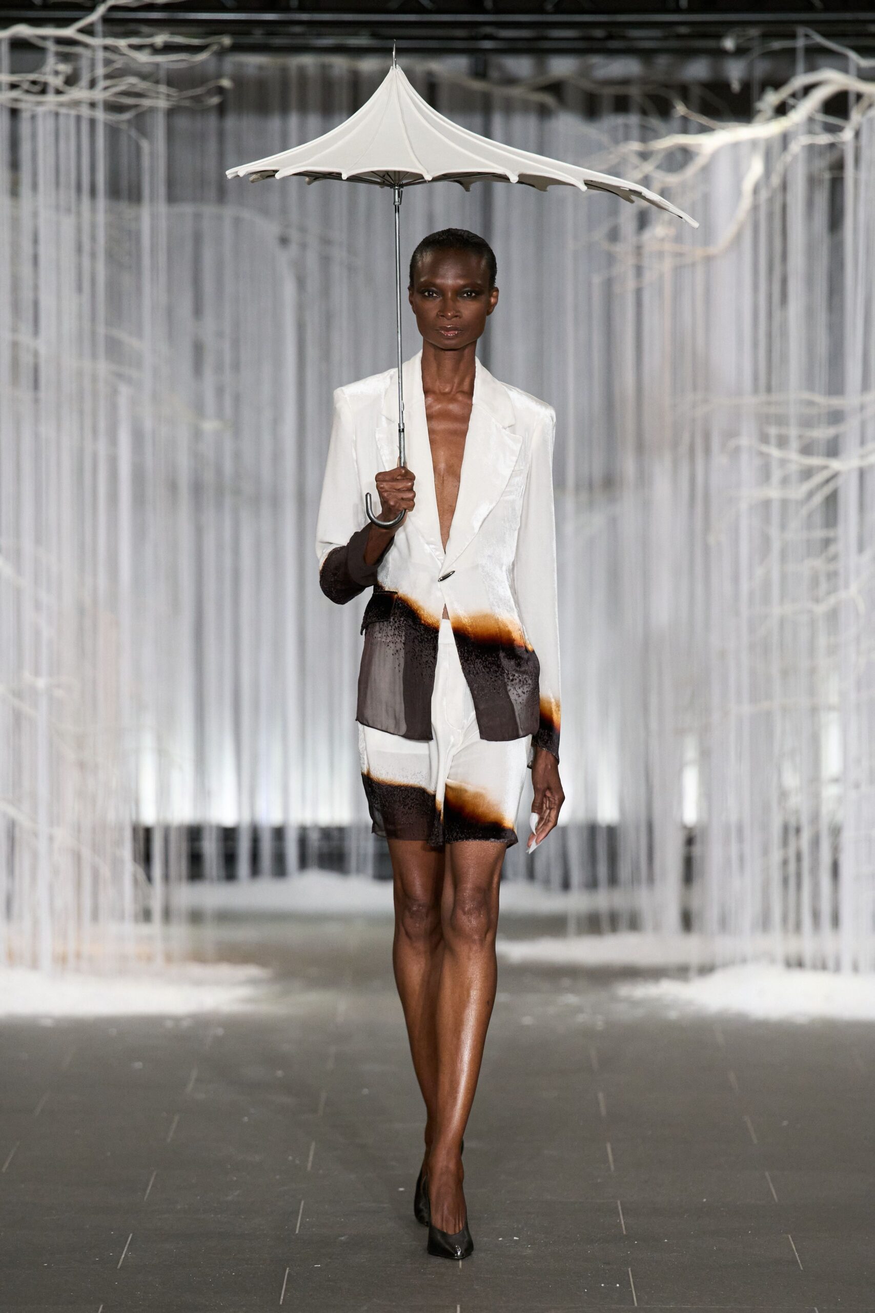 White pagoda umbrella with a sharply pointed finial and hooked handle. Deep-plunge ivory blazer, hem and cuff airbrushed to a singed brown fade. Matching sheer shorts with the same scorched bands above the hem. Black pointed pumps. Crossing a hotel forecourt in warm rain after the power flickered twice.