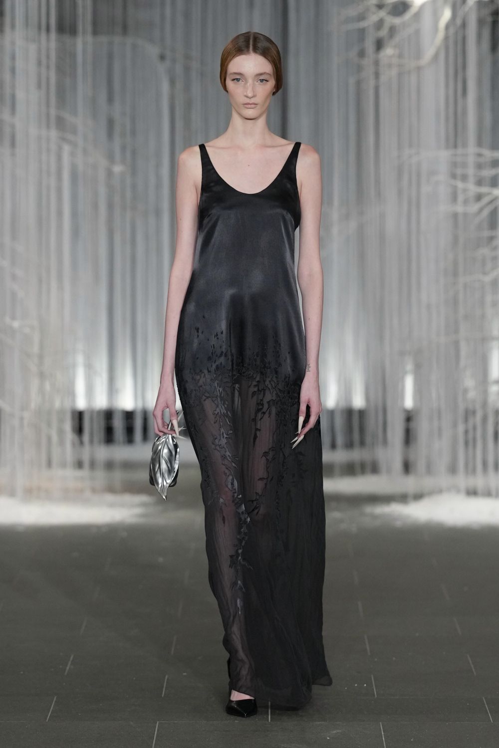 Black satin slip dress dissolving into a sheer devore skirt. Crushed silver pouch clutched in one hand. Black pointed pumps barely visible under the hem. Crossing a hotel atrium after midnight, fountain still running.
