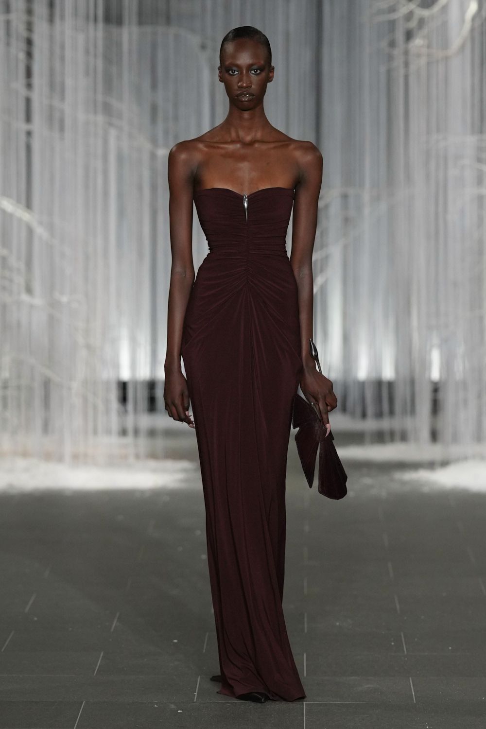 Strapless oxblood jersey gown, ruched inward to a sharp metal notch. Long silver cuff curling off the wrist like a blade. Soft matching clutch, folded into a narrow pointed shape. Black pointed toes just breaking the hem. Whatever you wear to cross a hotel lobby after midnight in rain.