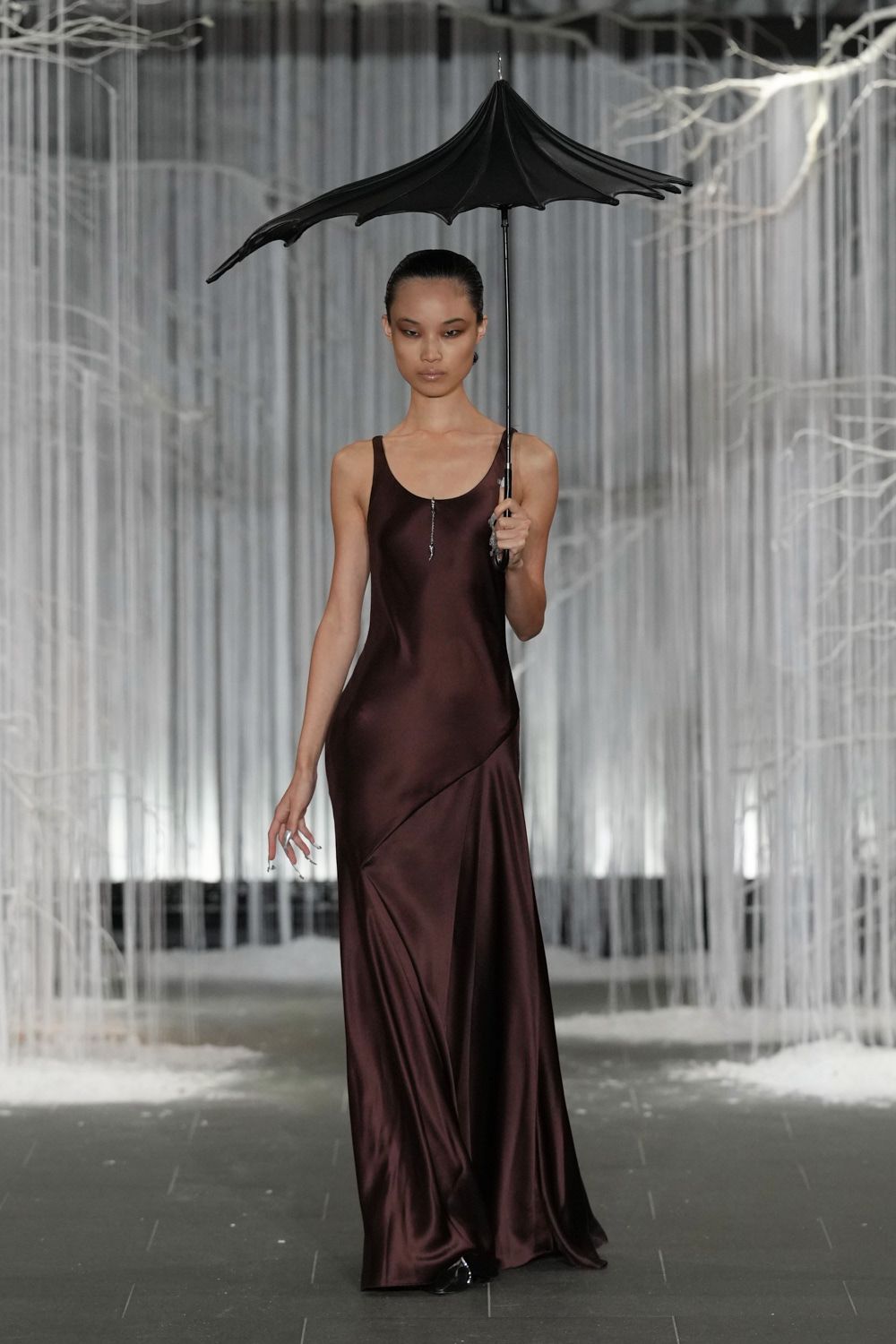 Black umbrella stretched into a batwing canopy with pointed ribs. Oxblood satin slip dress, bias-cut with a diagonal seam skimming the hip. Black pointed toes flashing under the pooled hem. Crossing a sculpture garden just as the first cold drops start.