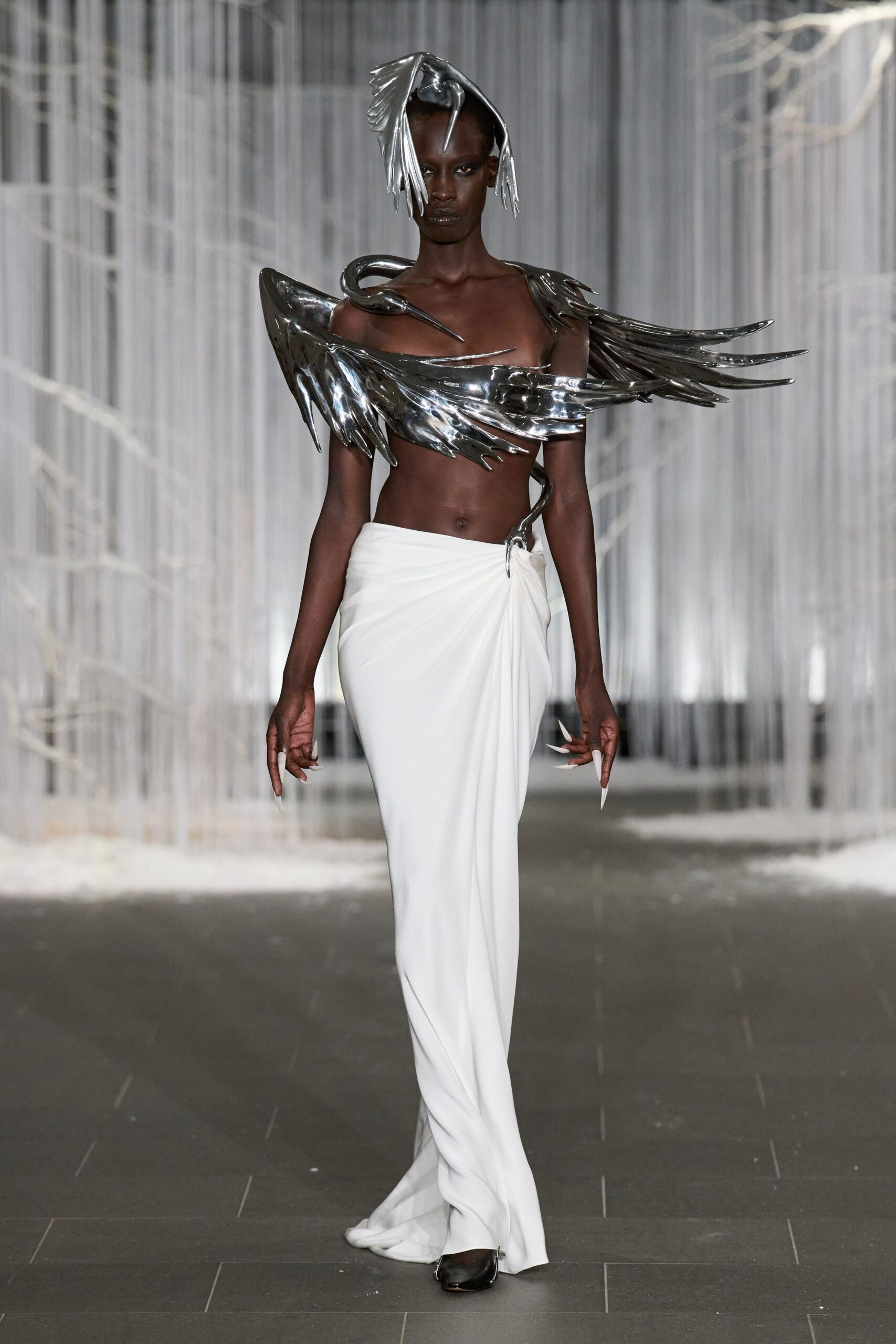 Molten silver bird hood, beak dropping over one brow. Mirror-metal wings circling the chest, one crane neck hooked at the collarbone. White draped column skirt, ruched into a sharp hip point. Long white talons. Black pointed shoes peeking under the hem. Crossing a frozen conservatory aisle after everyone else has gone home.