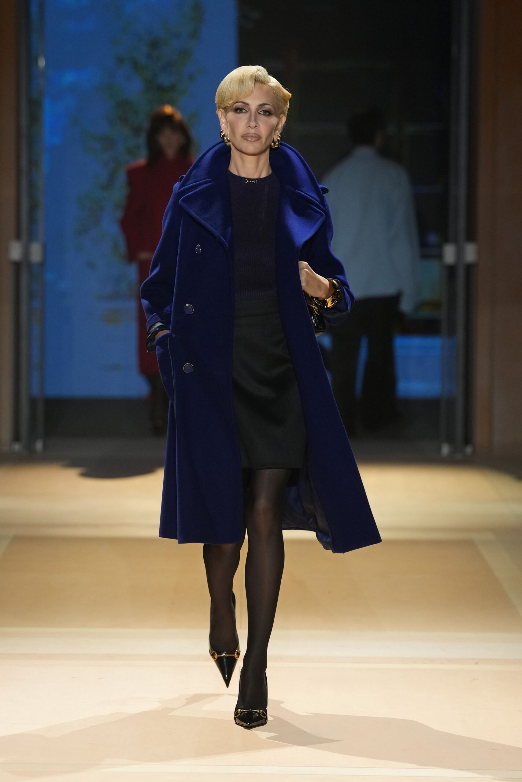 Cobalt wool coat with a huge sculpted shawl collar and dark buttons. Ink ribbed knit top, tiny gold bar closing the neckline. Black straight skirt cut high on the waist. Sheer black tights. Patent black pointed pumps with a gold horsebit. Chunky gold hoops. Crossing a hotel lobby before the bar crowd spills out.