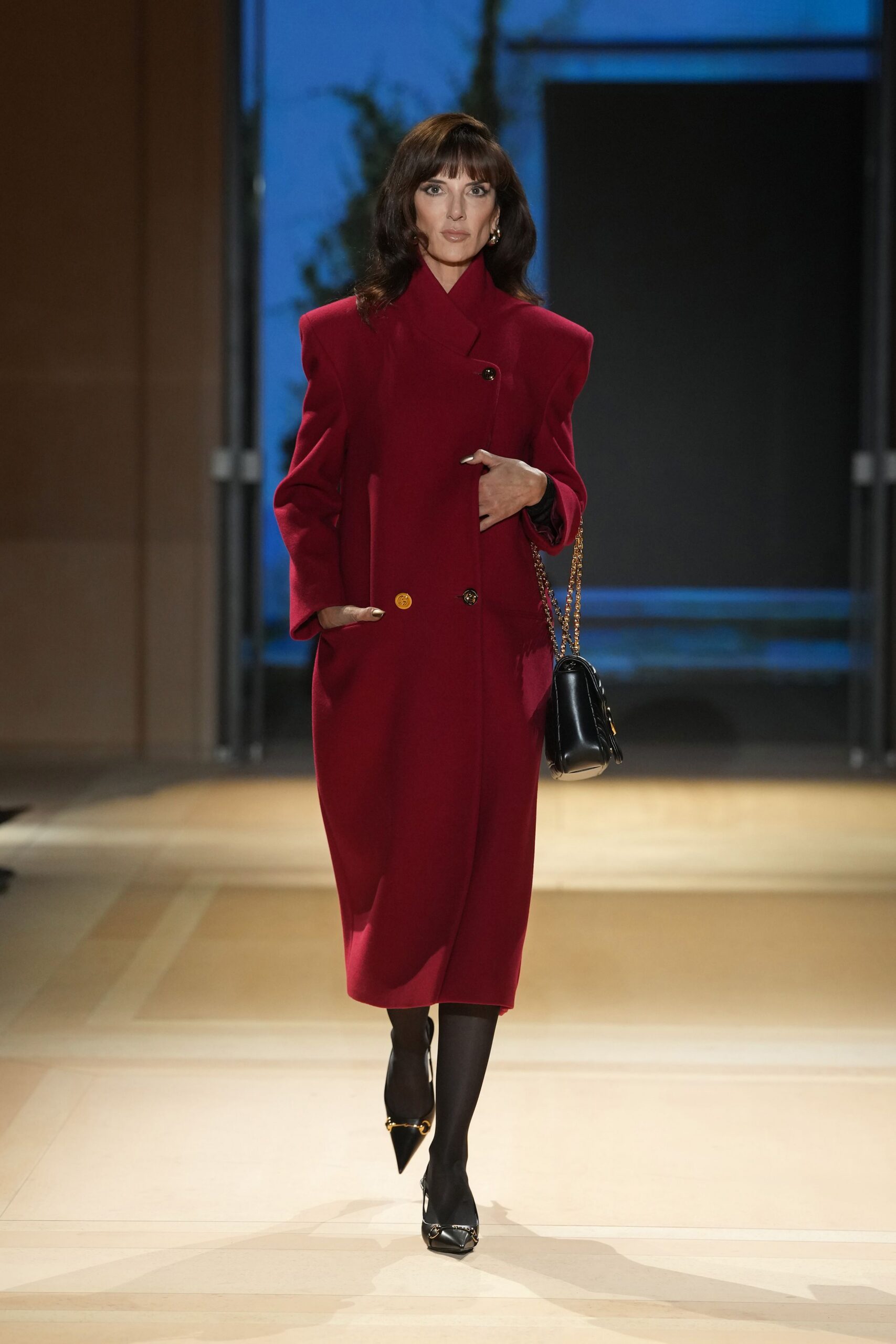 Cranberry wool coat with a wrapped stand collar and mixed buttons. Black leather shoulder bag on a fine gold chain. Opaque black tights. Black pointed pumps with horsebit hardware across the vamp. For crossing a hotel lobby before anyone else is downstairs.