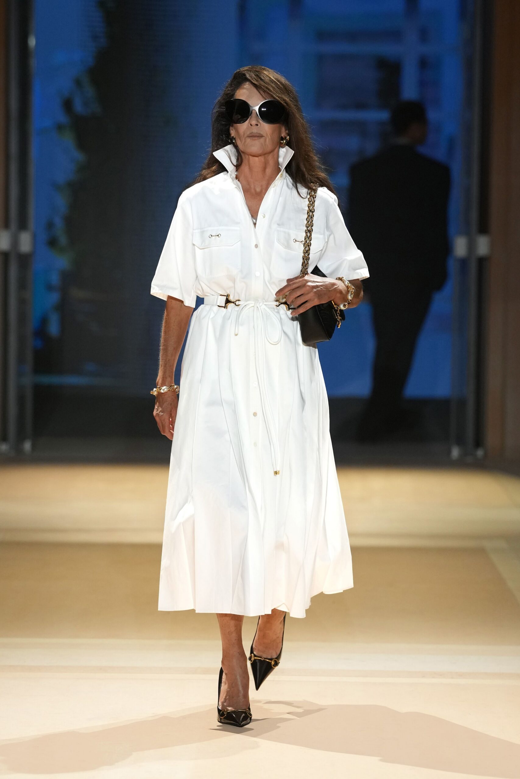 Oversized black sunglasses with thick ivory rims. White poplin shirt dress, collar flipped high, drawcord cinching the waist. Black leather shoulder bag on a chunky gilt chain. Black patent pumps with horsebit hardware across the pointed toe. Hotel atrium at dusk, keys in hand before asking for an upgrade.