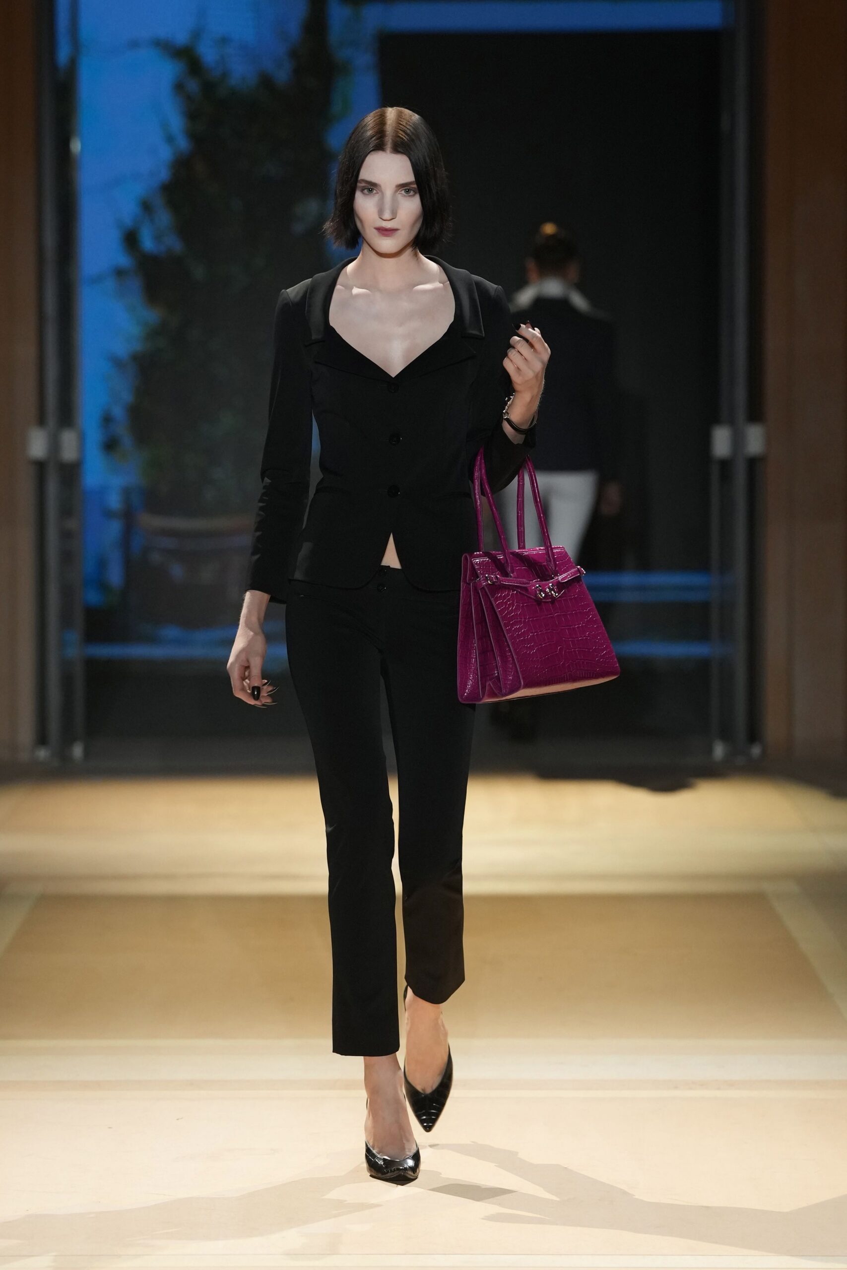 Black tailored blazer with a deep sweetheart opening and single low vent. Black slim trousers breaking lightly above the ankle. Magenta crocodile-embossed tote with silver turn-lock straps. Black patent pumps with sharply pointed toes. Crossing a hotel lobby before anyone else is down for breakfast.