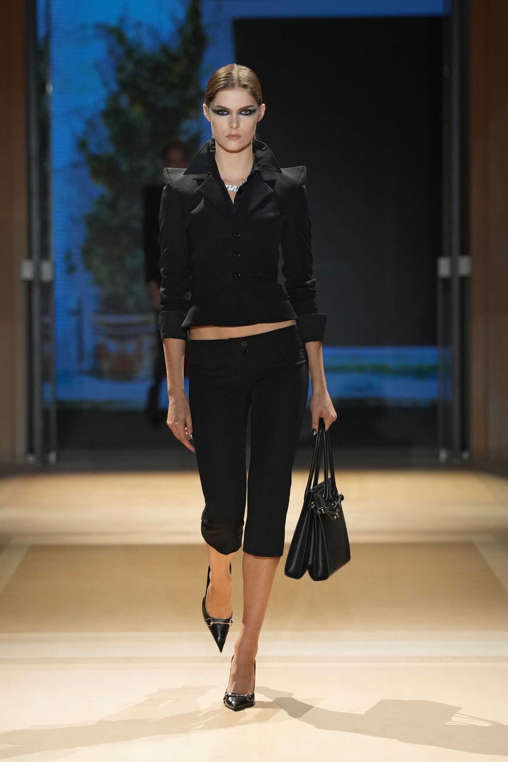 Black fitted jacket with peaked shoulders and a raised collar. Low-slung black capris, the third button catching the light. Short silver necklace at the open throat. Black leather tote with doubled shoulder straps. Black pointed pumps with a small metal bit. For crossing a hotel lobby before anyone else is downstairs.