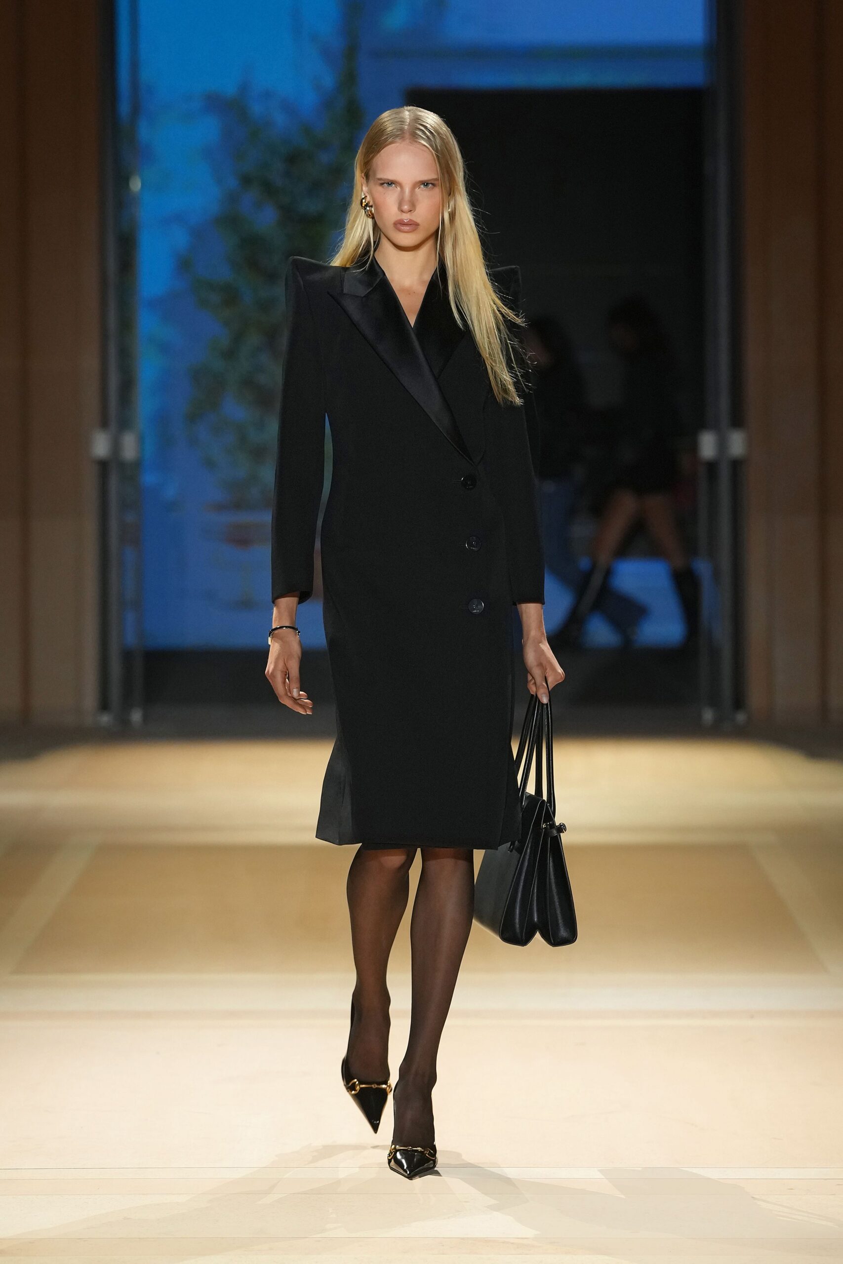 Black wool coatdress with satin shawl lapels and hard peaked shoulders. Slim black bangle at one wrist. Sheer black tights. Black double-gusset tote with long thin handles. Patent black pumps with a gold horsebit across the pointed toe. For the first dinner after the divorce papers are signed.