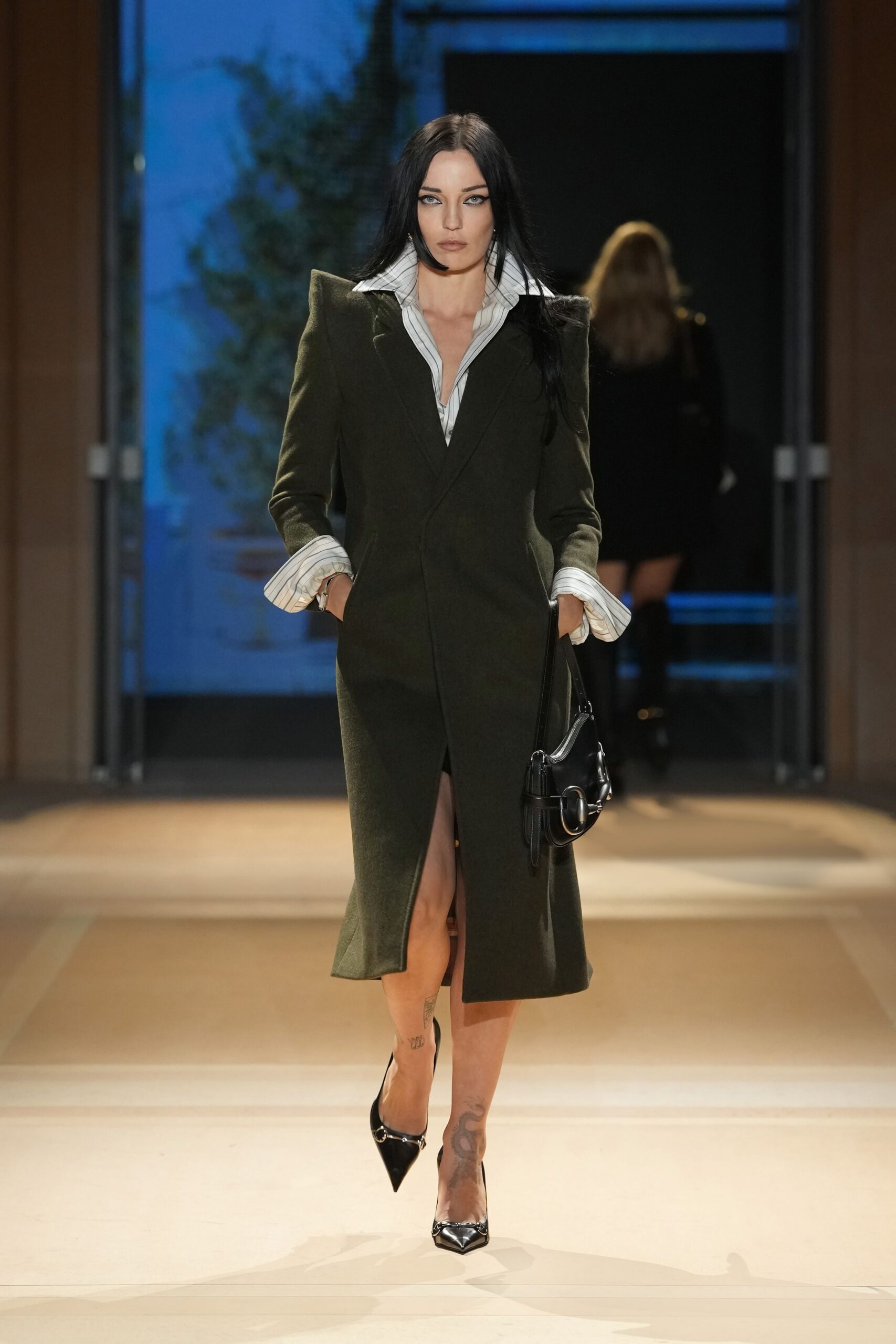 Dark moss wool coatdress with sharp padded shoulders and a front slit. White pinstriped shirt, collar flared high and cuffs turned back wide. Black leather shoulder bag with oversized silver ring hardware. Black patent pointed pumps capped with curved silver metal. Walking into a hotel bar before the rain has soaked your hair.