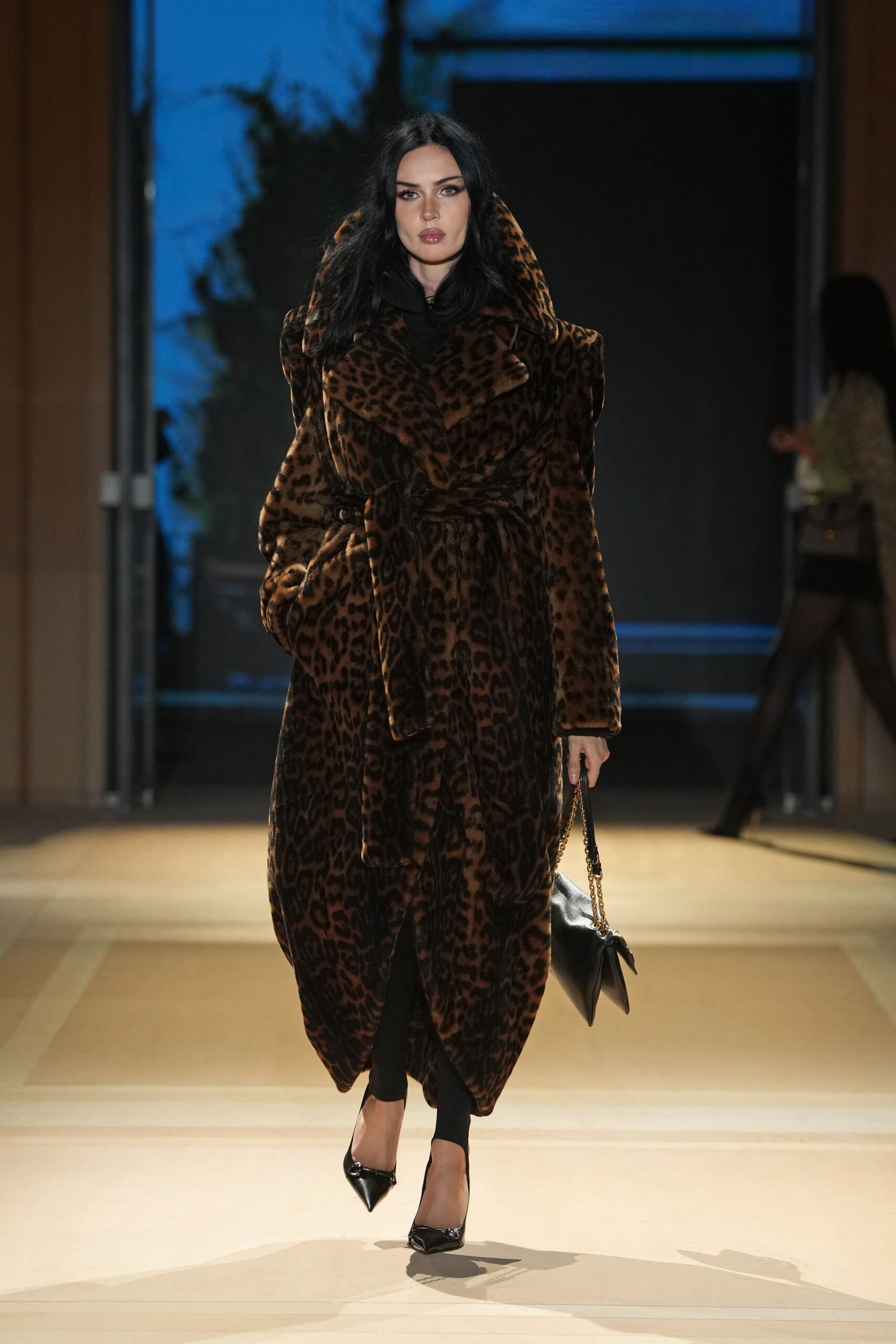 Long leopard faux-fur wrap coat, peaked shoulders and a thick self-belt knot. Black stirrup leggings disappearing under the coat hem. Black leather bag on a gold chain strap. Black pointed pumps with a small knotted bow at the toe. Hotel lobby after midnight, still carrying the room key card.