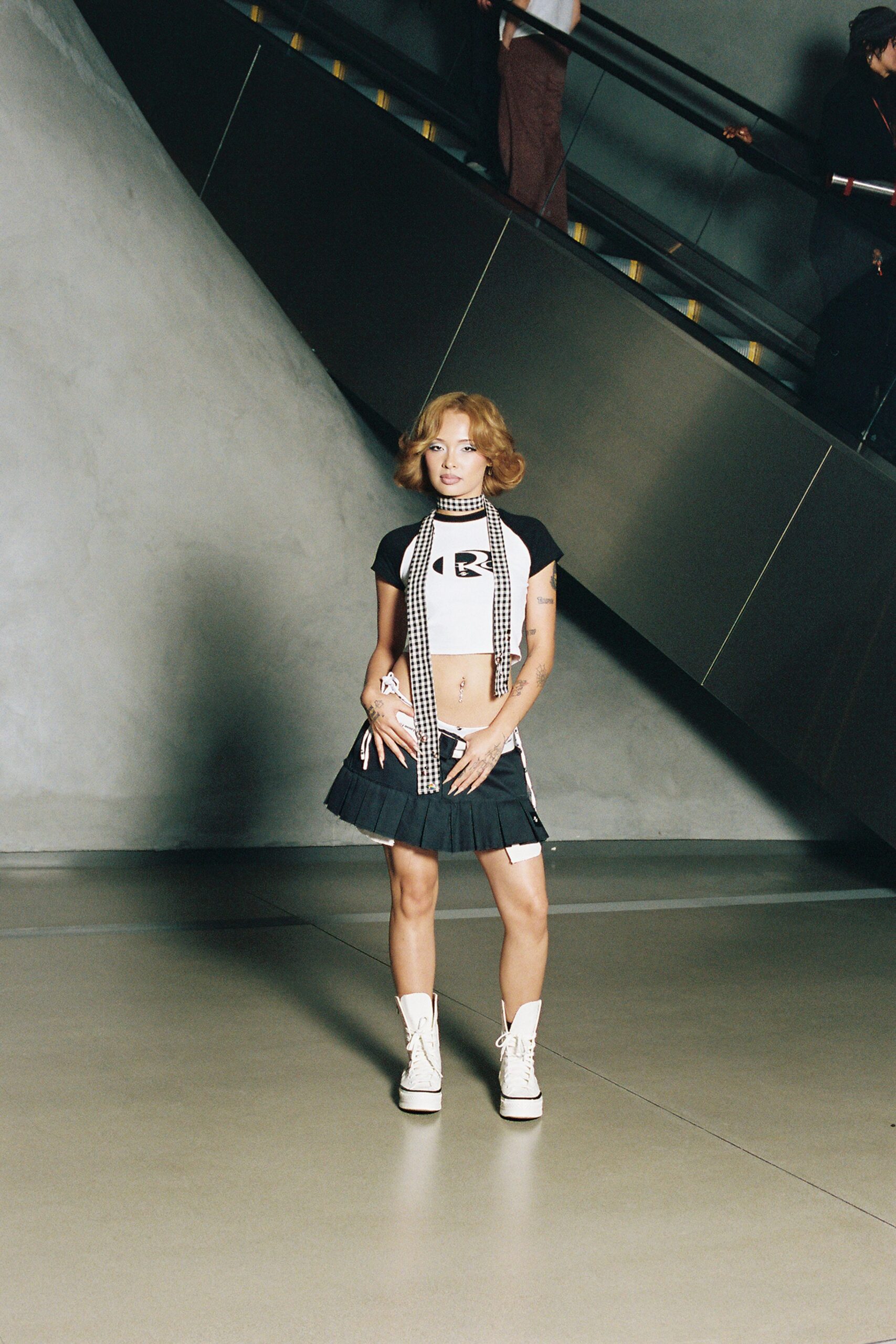 White stretch crop top with black raglan sleeves and a chest graphic. Long black-and-white gingham scarf looped tight as a choker. Black pleated mini riding low, one white tie hanging loose. White platform lace-up boots with rubber foxing at the sole. Riding the mall escalator after buying nothing but a sugary drink.