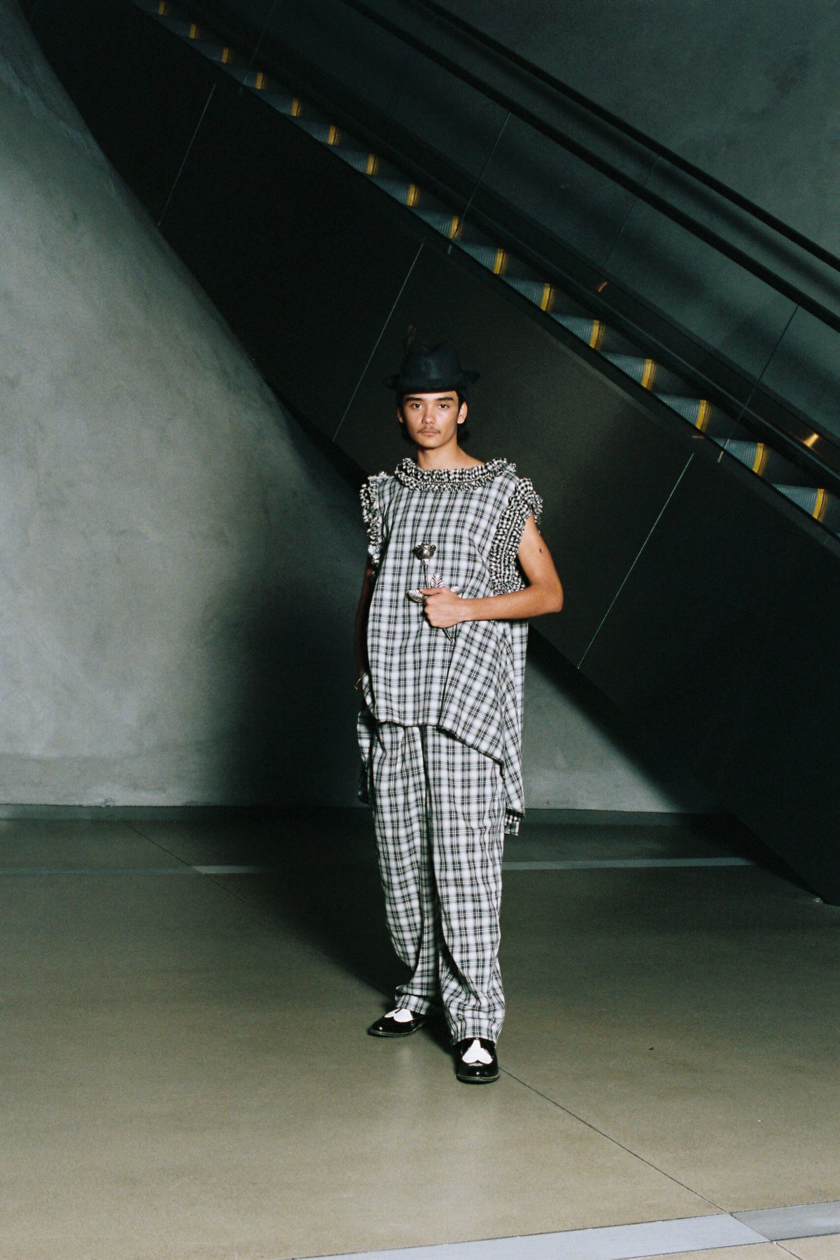 Black felt trilby with a pinched crown and narrow brim. Long black-and-white plaid tunic, neckline and armholes ringed in ruffled checks. Matching plaid trousers falling loose to a slight break at the shoe. Black-and-white spectator flats, glossy leather. Crossing the museum atrium before the escalators have started moving.