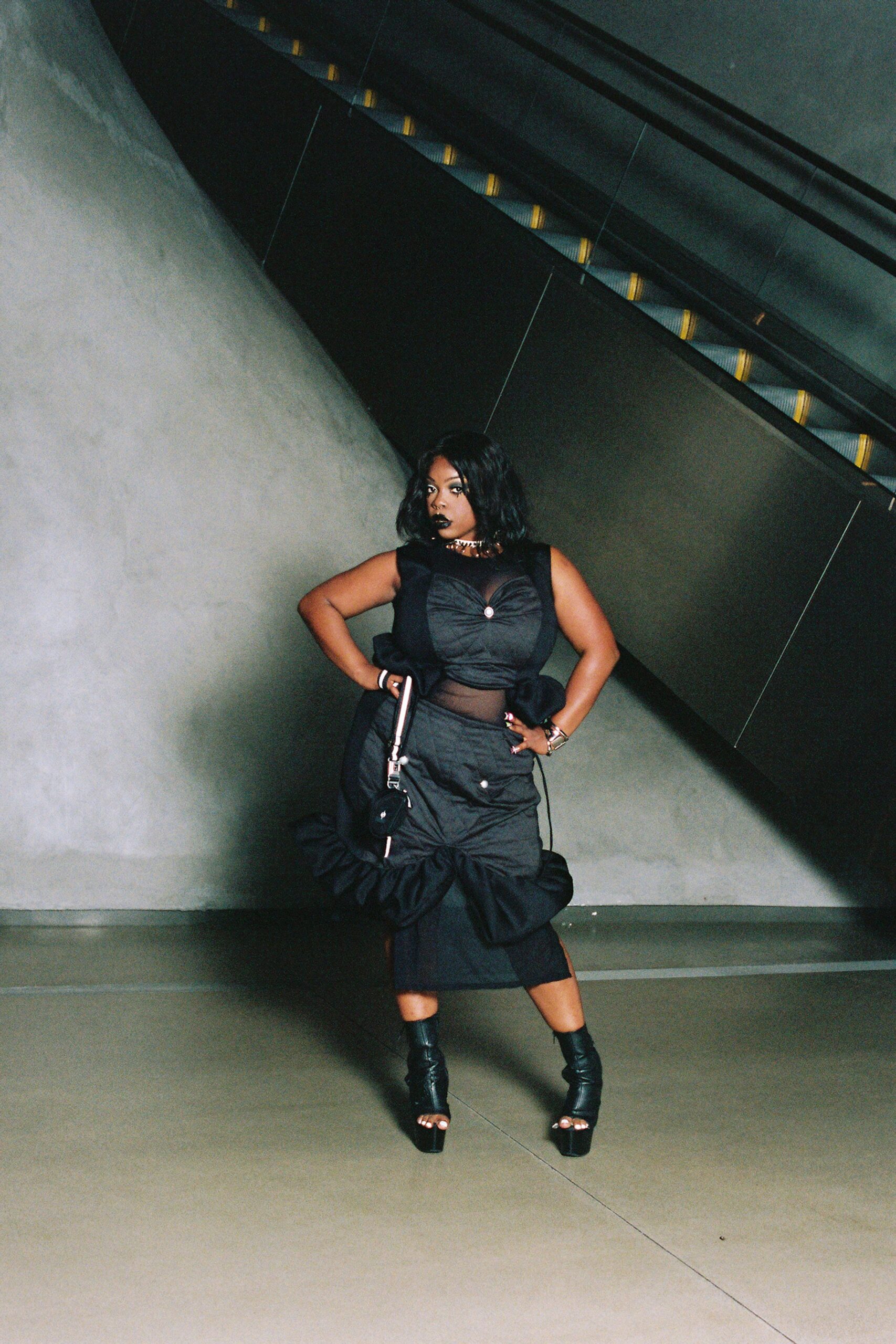 Black ruched dress with sheer mesh midriff and oversized ruffles at the hips. Small black shoulder bag with a pale strap and dangling buckle tab. Black peep-toe platform sandals, the front slit opening around bare toes. Chunky silver choker close at the throat. Waiting under an escalator after missing the last museum talk.