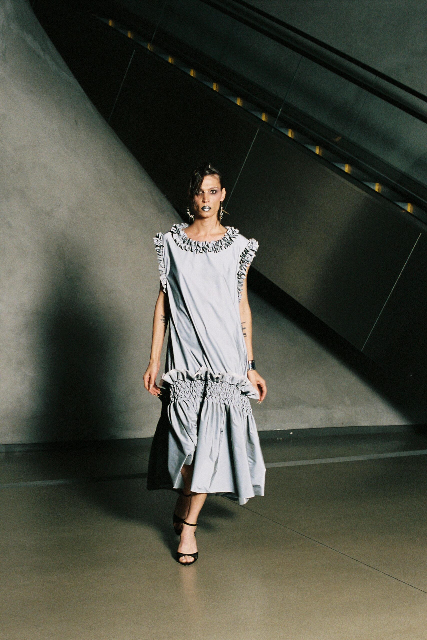 Pale grey shift dress with black-edged ruffles at neck and armholes. Dropped bubble hem caught into dense shirring above the calf. Black ankle-strap sandals, narrow front band over bare toes. Mismatched dangling silver earrings. Crossing an empty transit hall after the last moving walkway stopped.