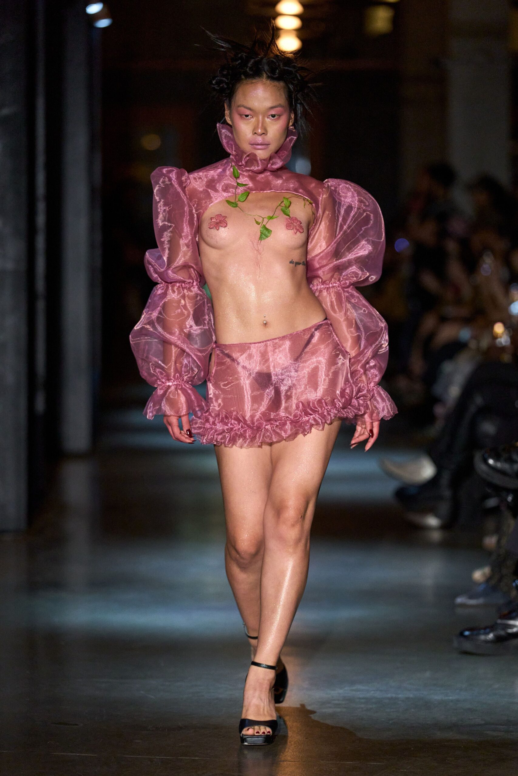 Rose organza cutout top with a wired ruff and balloon sleeves. Real green vine trailing diagonally across the bare chest. Matching organza mini with a dropped waist and ruched bubble hem. Black ankle-strap platform sandals. Walking home from a costume party at dawn, still glittered with sweat.