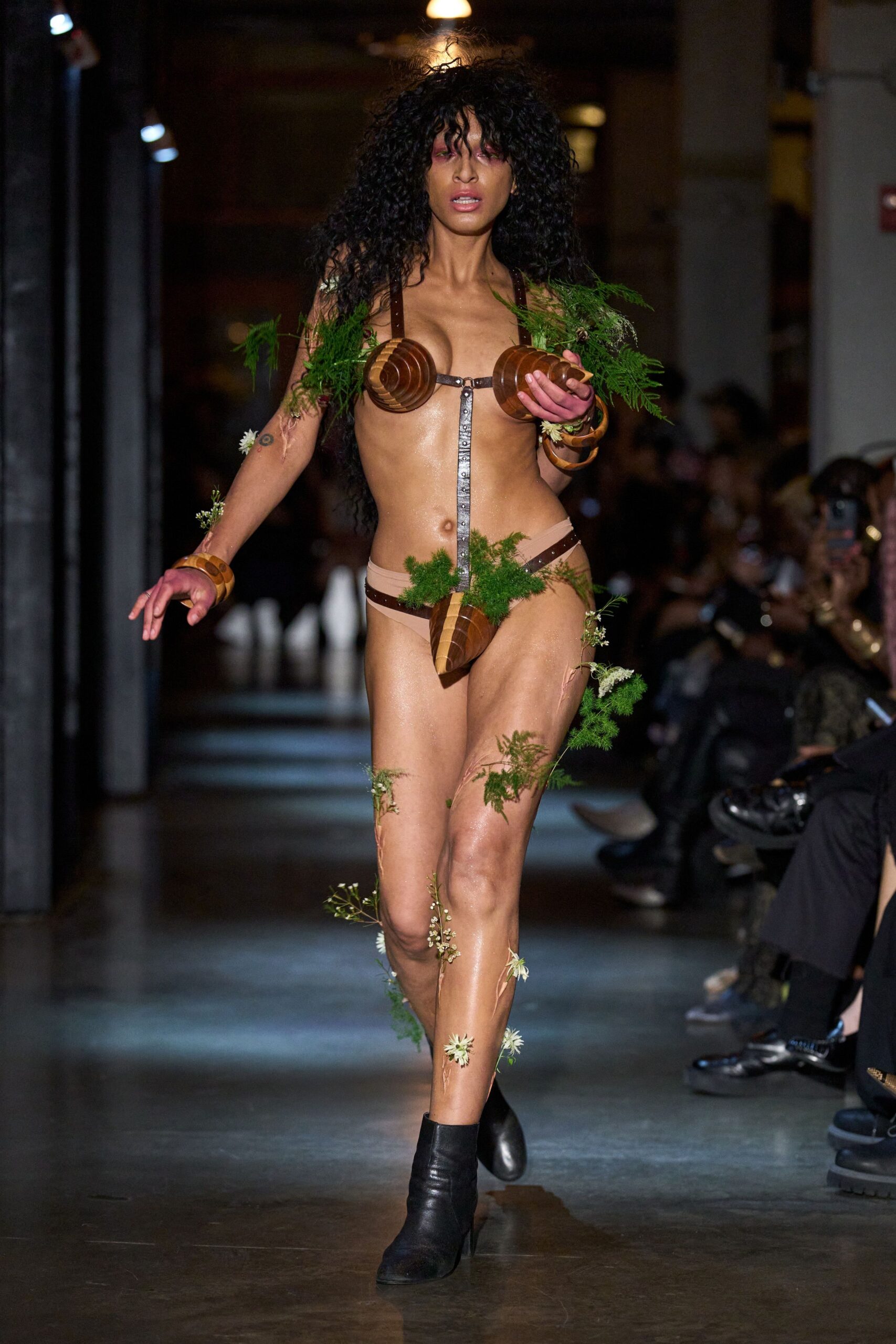Harness bra with turned wooden cone cups and fern sprigs. Nude thong slung from brown studded straps, wooden codpiece centered low. Stacked carved wooden bangles at both wrists. Black leather ankle boots with a blunt squared toe. For slipping out of a pagan fair after midnight rain.