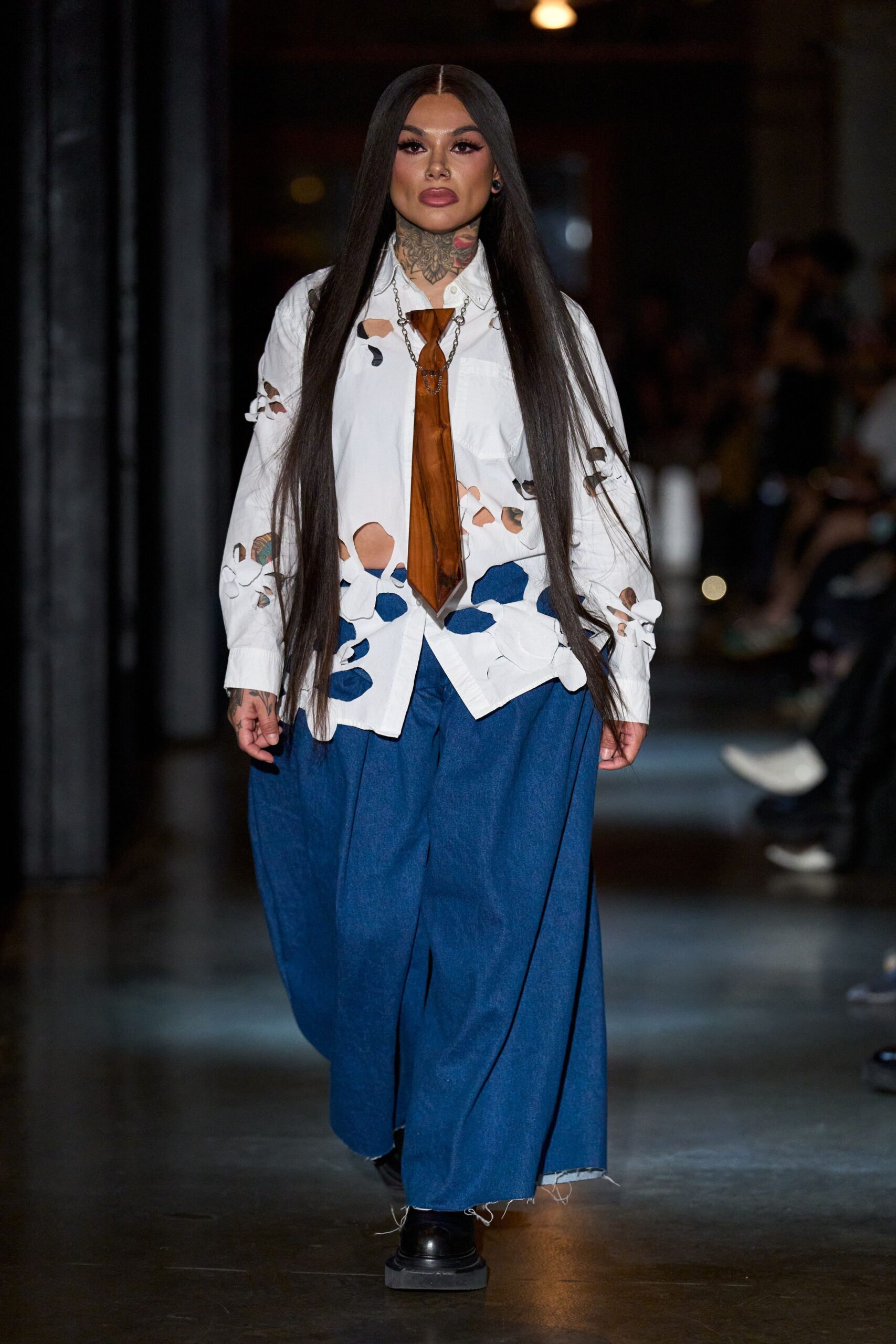 White button-down slashed with irregular cutouts and tied sleeve openings. Burnt-caramel satin tie hanging narrow under a silver chain loop. Indigo wide-leg trousers with deep pleats and frayed hems. Black square-toe shoes. Whatever you wear to read tarot in a warehouse after rain.