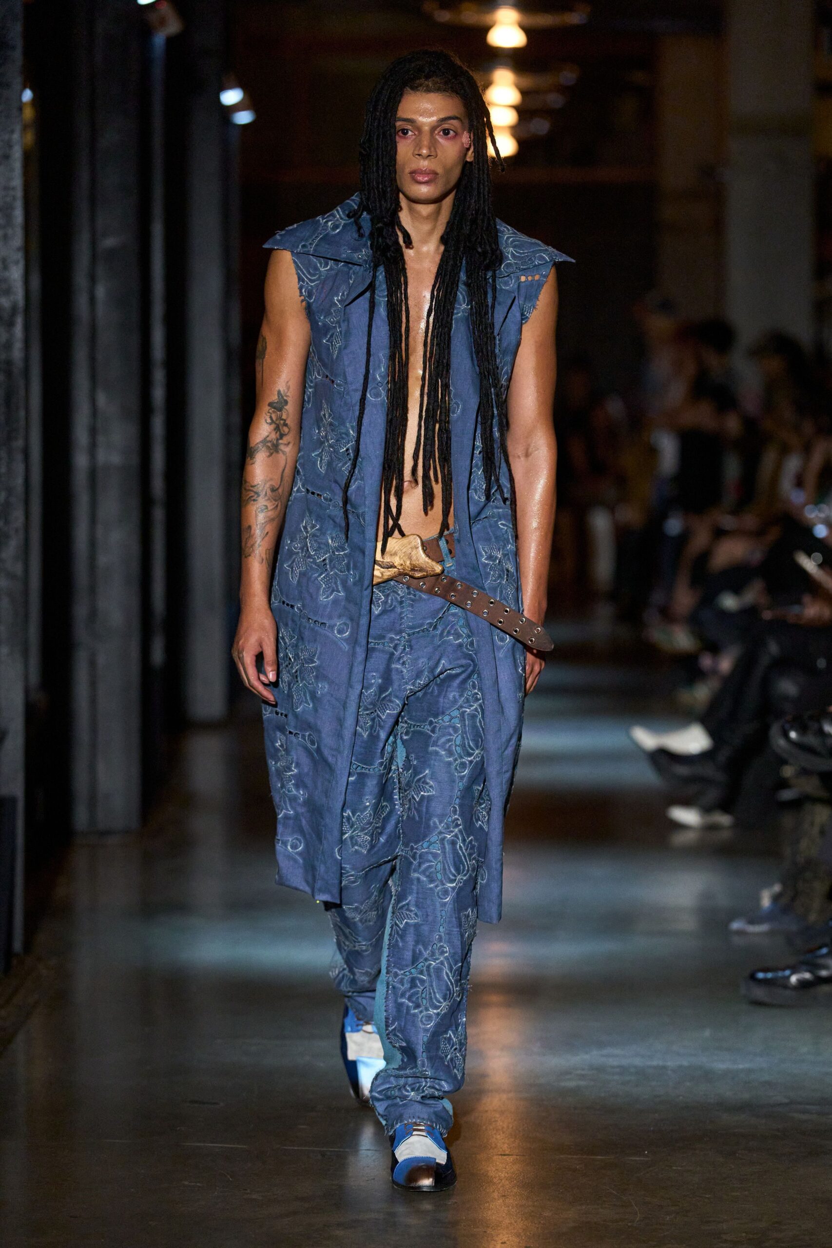 Dusty blue sleeveless coat with broad folded shoulders and raw armholes. Matching loose trousers in stitched floral jacquard, hem pooling over the shoe. Small pale hide pouch slung low on a grommeted brown belt. Blue and white sneakers with a dark capped toe. For crossing the station concourse after missing the last easy train.