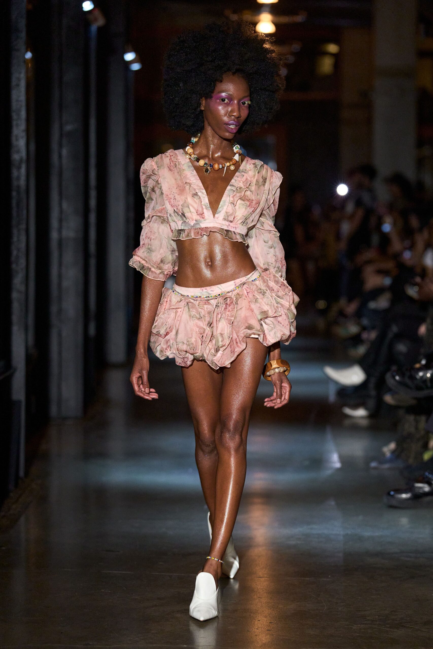 Blush rose-print chiffon crop top with ruffled plunge and puffed sleeves. Matching bubble shorts, gathered into petal-like ruffles at the hips. Chunky bead necklace with a small tooth-shaped drop. Thick amber bangle. White square-toe heels. Walking back from a beach bar before the salt dries.